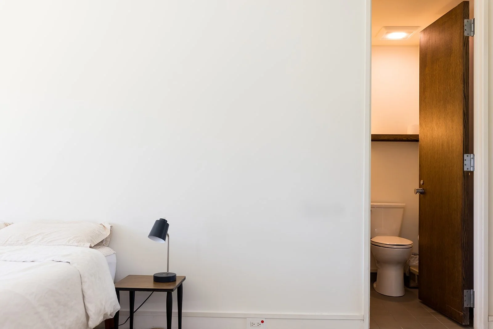 Minimalist bedroom with bed, pillows, bedside lamp, and a peek into a small attached bathroom.