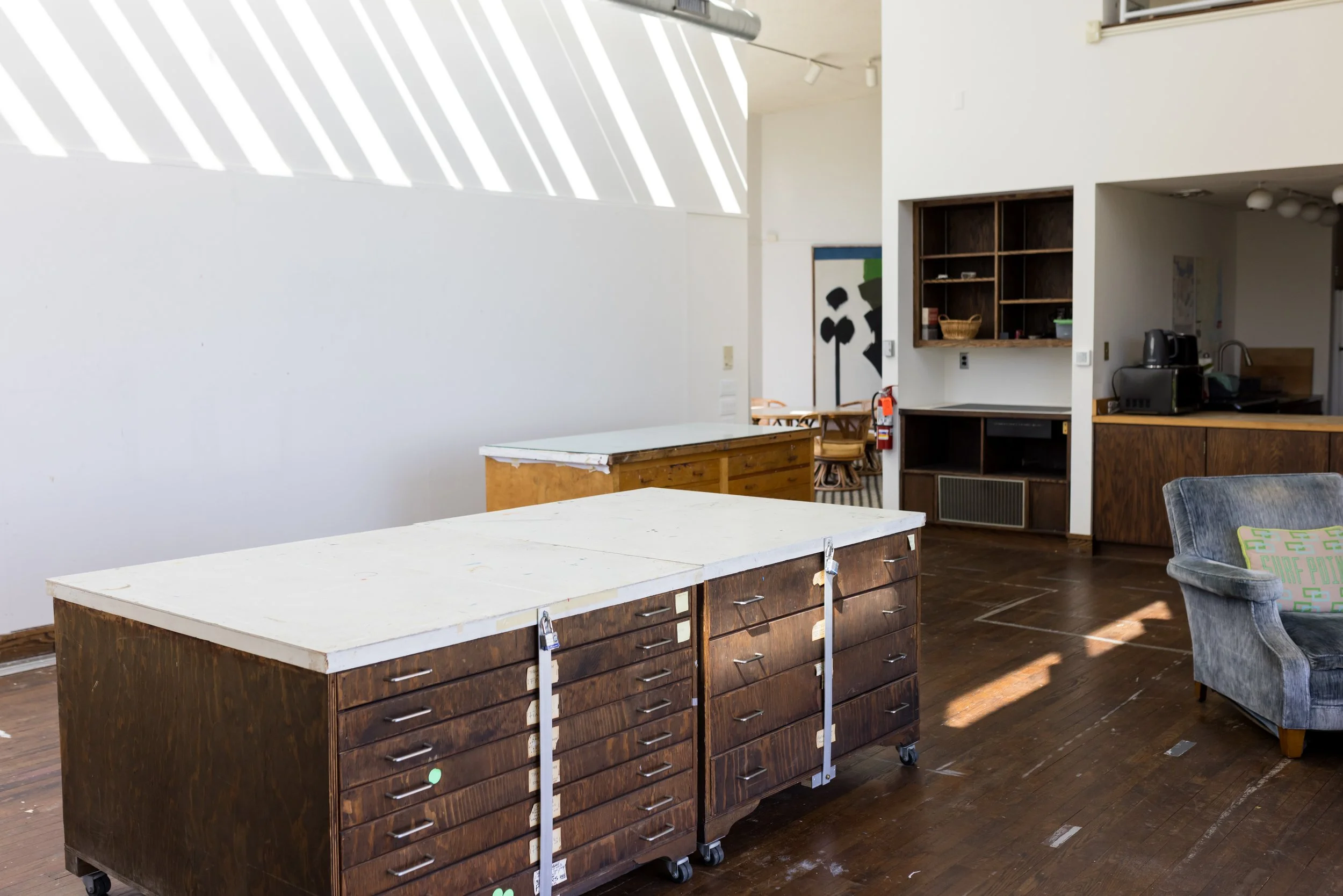 Interior of an art studio with large tables, storage cabinets, a gray armchair, and shelving. Sunlight streams through windows creating shadows on white walls and a wooden floor. A kitchenette area is partially visible in the background.