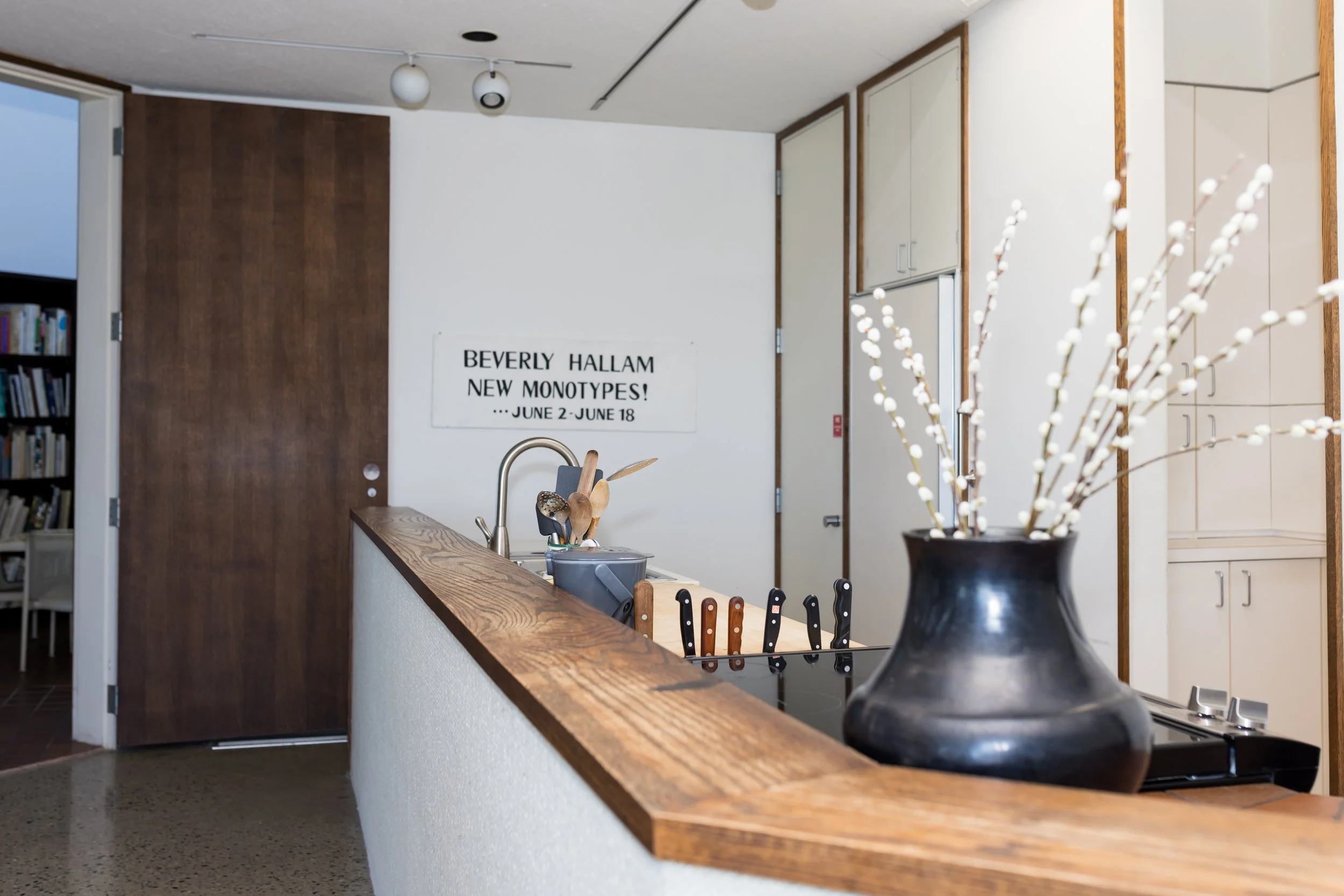 Modern kitchen with wooden countertop, black vase with white branches, knives on a magnetic strip, wooden utensils in a pot, and a poster on the wall mentioning an art exhibition by Beverly Hallam.