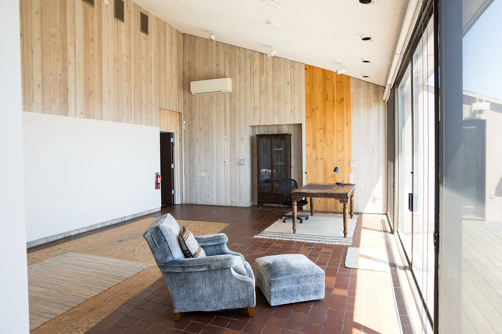 Interior of a minimalist room with wooden walls and brown tiled floor, featuring a rustic wooden table, and an arm chair and ottoman. Large windows offer a view of a porch and the ocean outside.