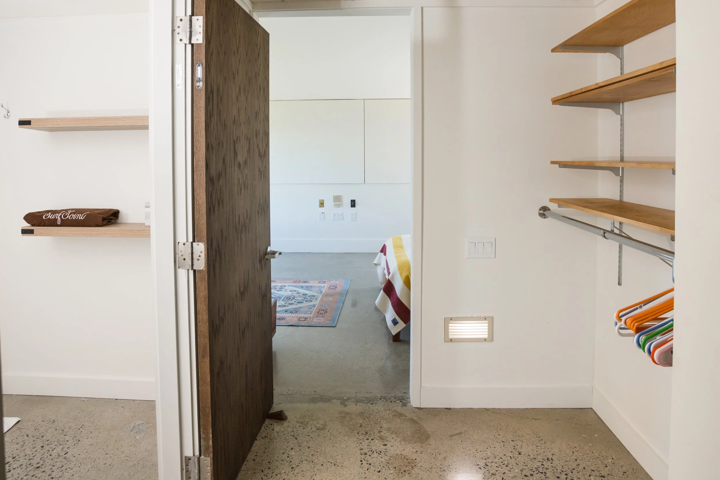 Open door leading from a closet area into a bedroom with a colorful rug and bed. The closet has wooden shelves, a hanging rod with clothes hangers, and a neatly folded dark brown towel on a shelf. The floor is polished concrete.