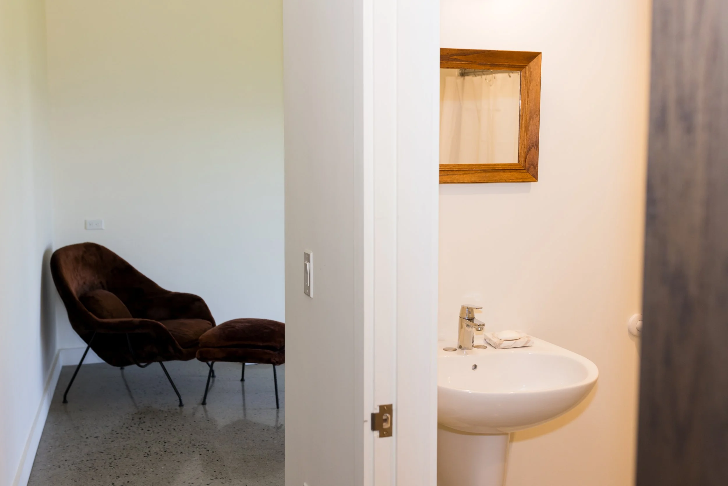 Modern interior with a brown upholstered chair, matching footrest, white walls, a narrow doorway leading to a bathroom with a pedestal sink, and a framed mirror on the wall.