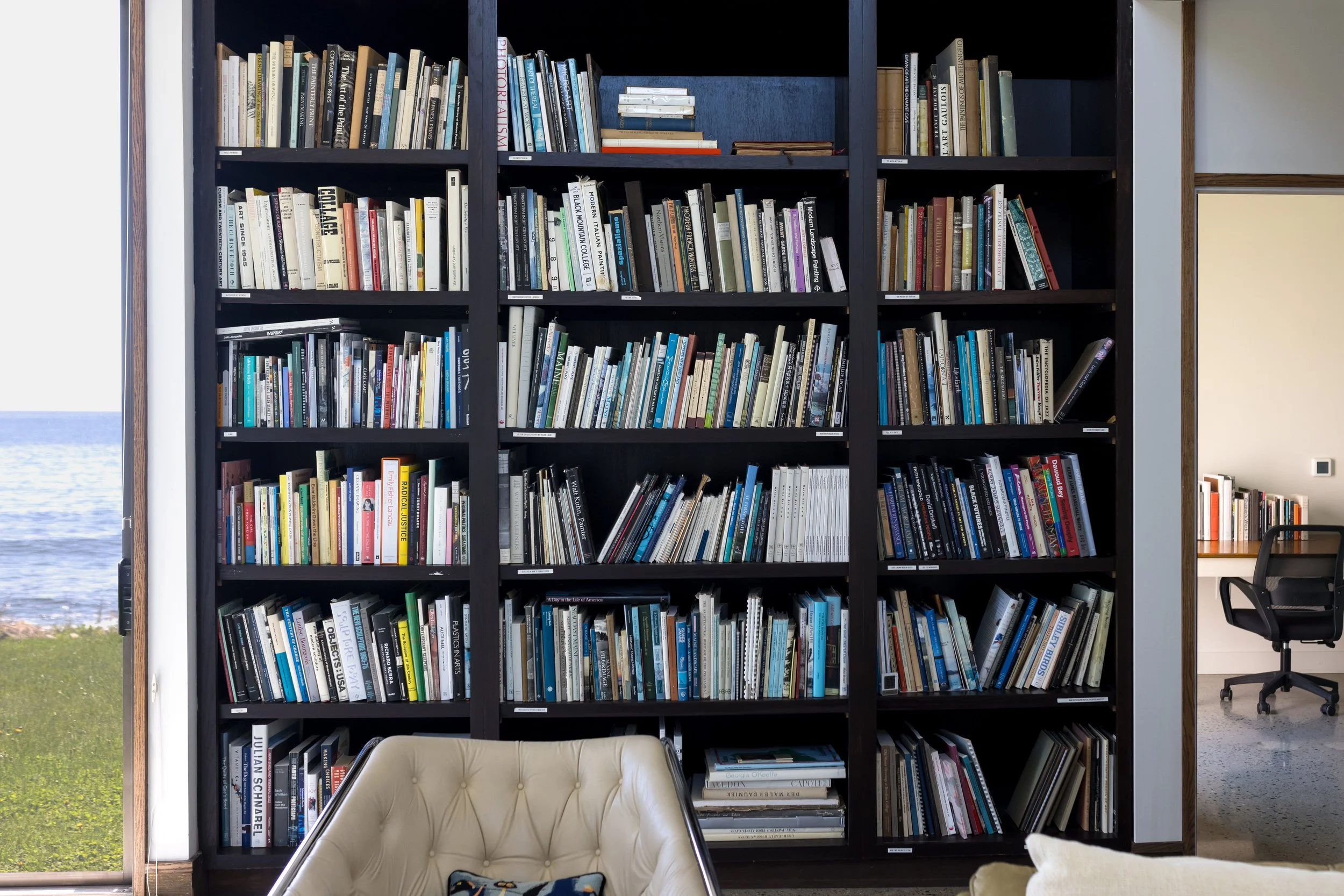Bookshelf filled with books, desk and chair, ocean view through window.