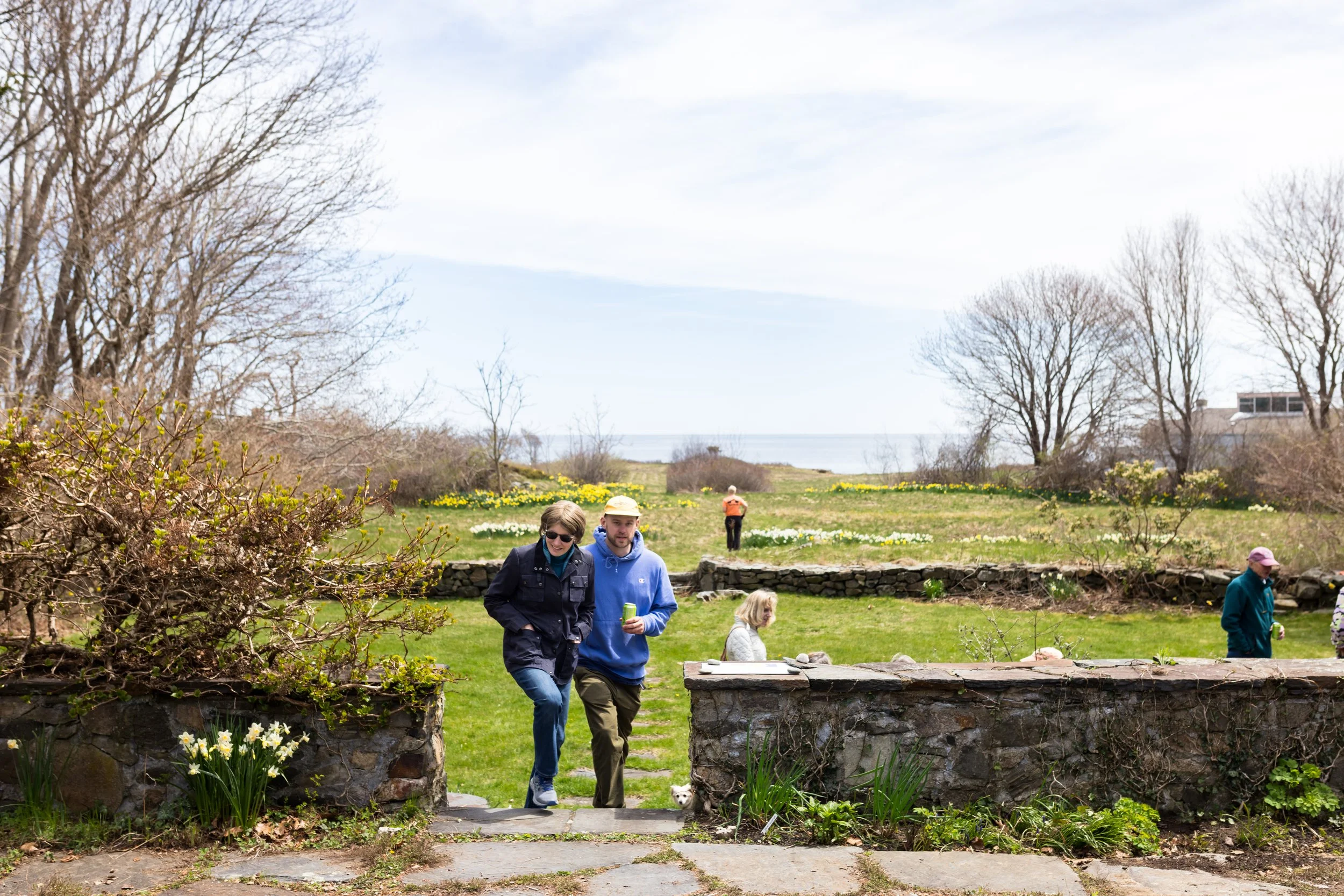 People walking in a grassy garden with trees and ocean in the background, featuring stone walls and blooming flowers.