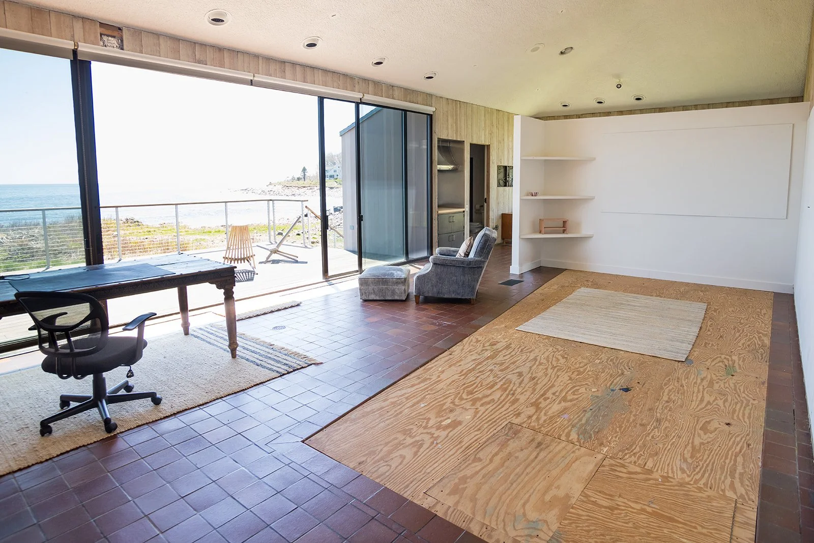 Interior of a minimalist room with wooden walls and brown tiled floor with a plywood section, and a half wall dividing the studio from the bedroom. It features a rustic wooden table and a desk chair, an arm chair and ottoman. Large windows offer a vi
