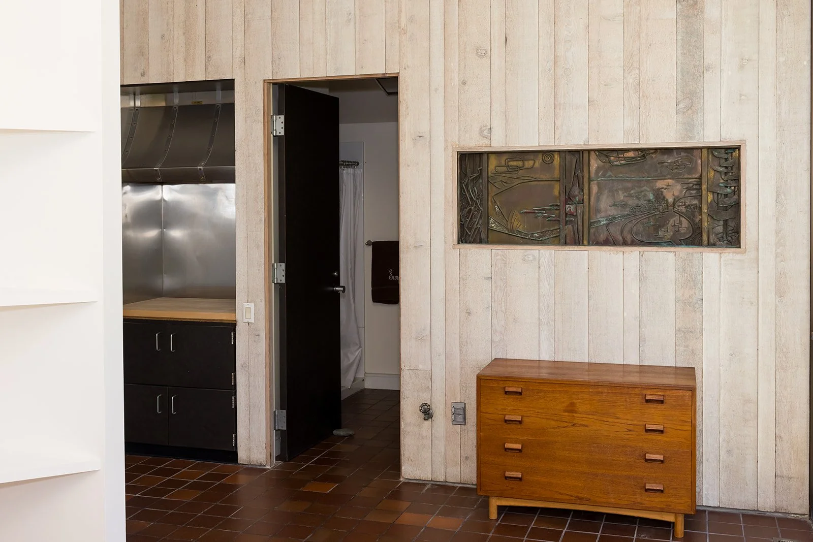Interior of a minimalist room with wooden walls and brown tiled floor, view from bedroom looking at a small wooden dresser, cabinets, artwork, and a peek into a bathroom.