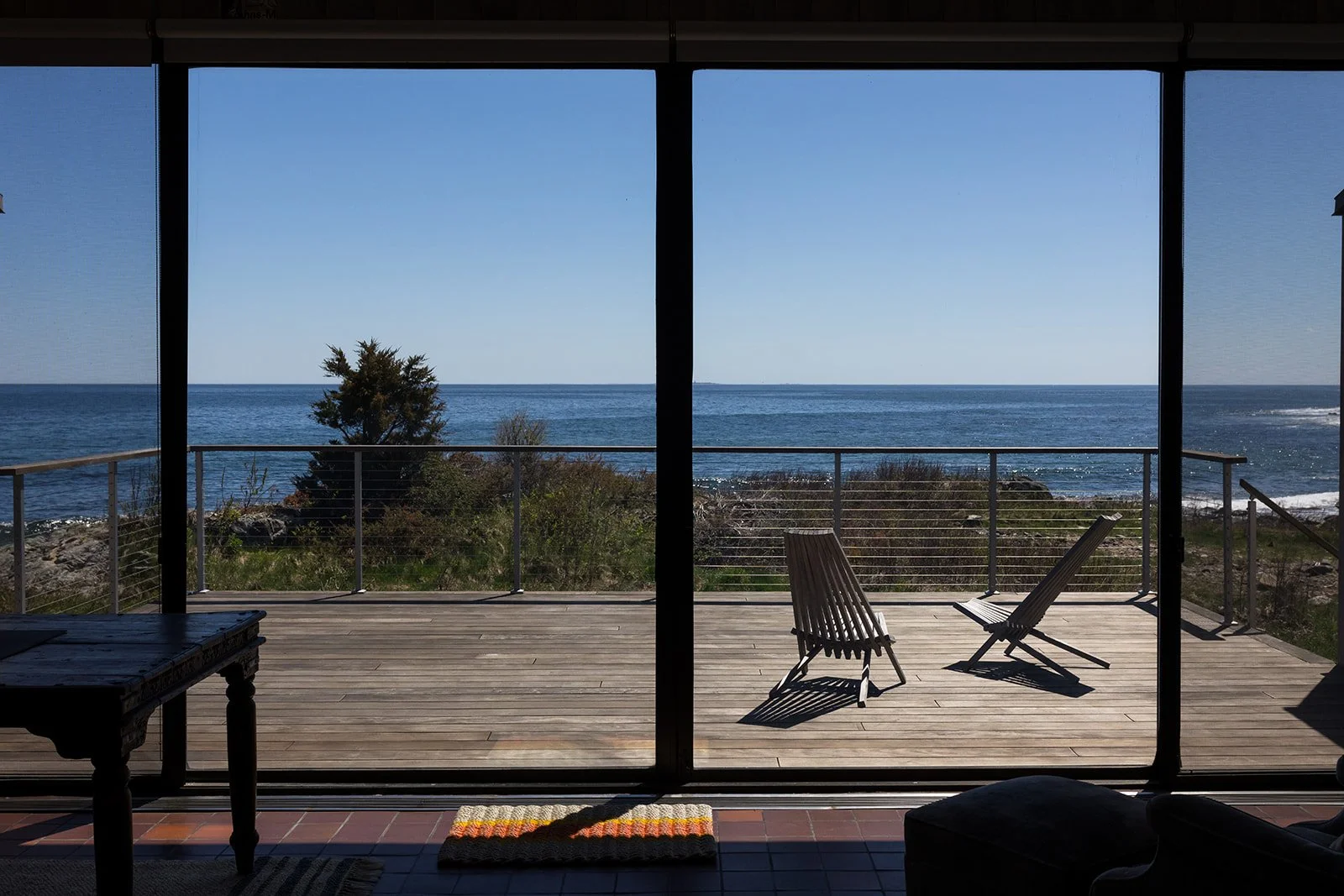 Interior of a minimalist room looking through large windows that offer a view of a porch with lawn chairs, and the ocean and trees outside.