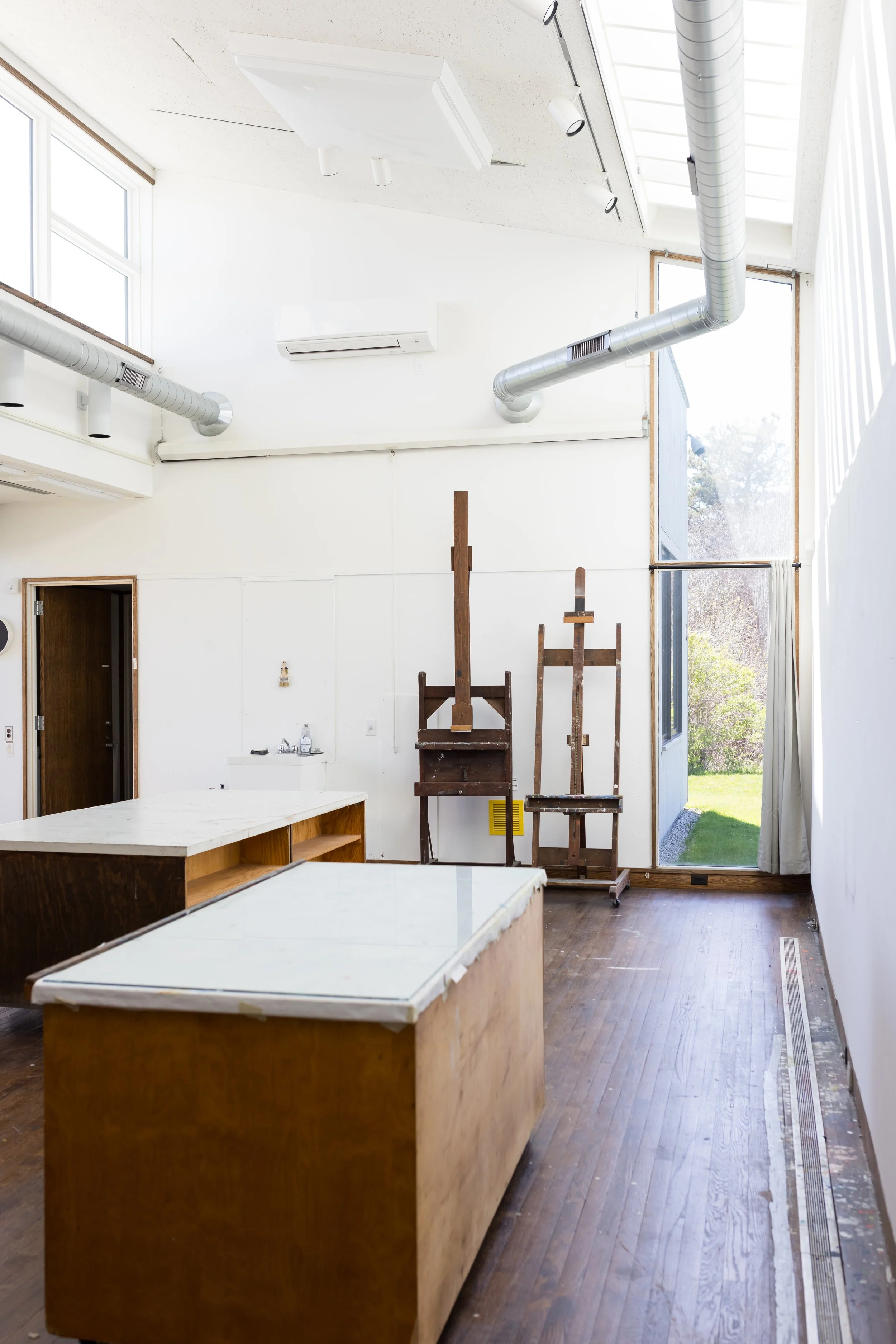 An art studio with wooden easels, large tables, white walls, high ceilings with skylights, ductwork, and a wooden floor. A door opens to a green lawn outside.