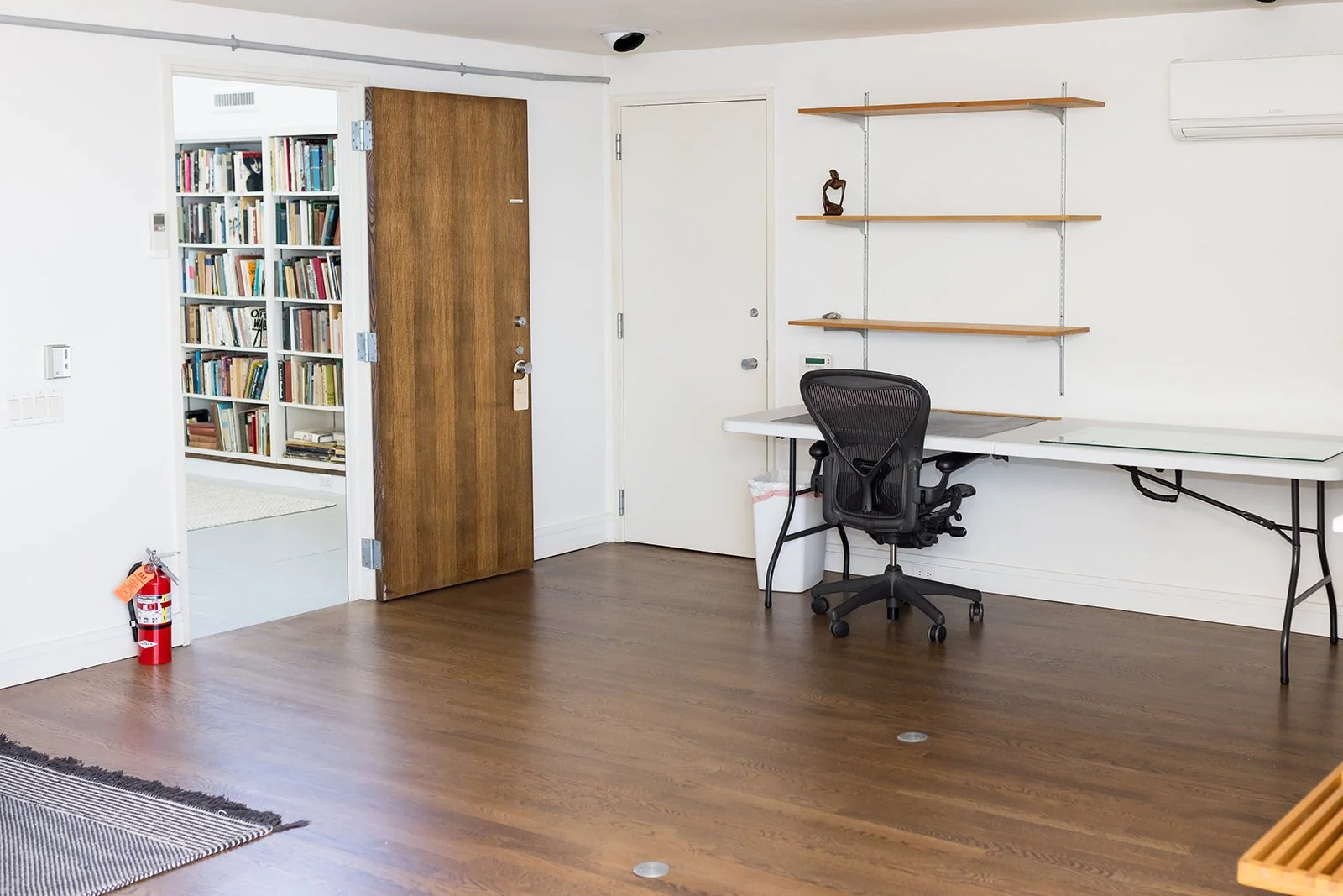 Minimalist room interior with wooden floor, one black desk chair, a folding white table, wall mounted shelving, and a door opening into a small library on an upper loft level.