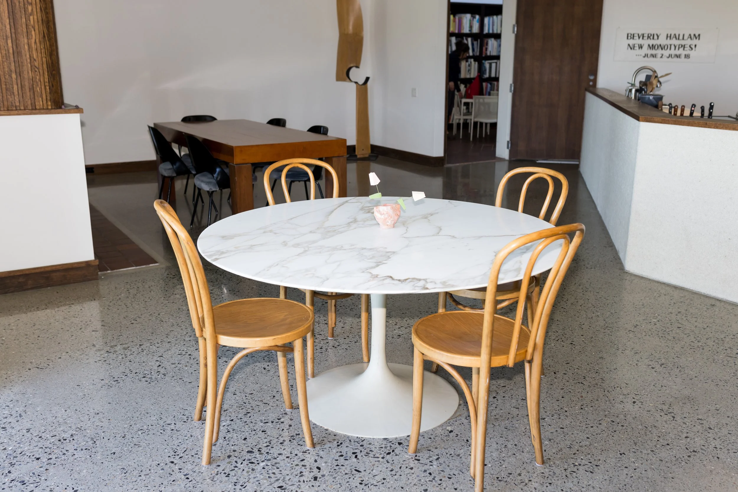 Modern interior with marble round table and wooden chairs, polished concrete floor, and a wooden bench in the background. Art sign on wall, with bookshelves visible in adjacent room.