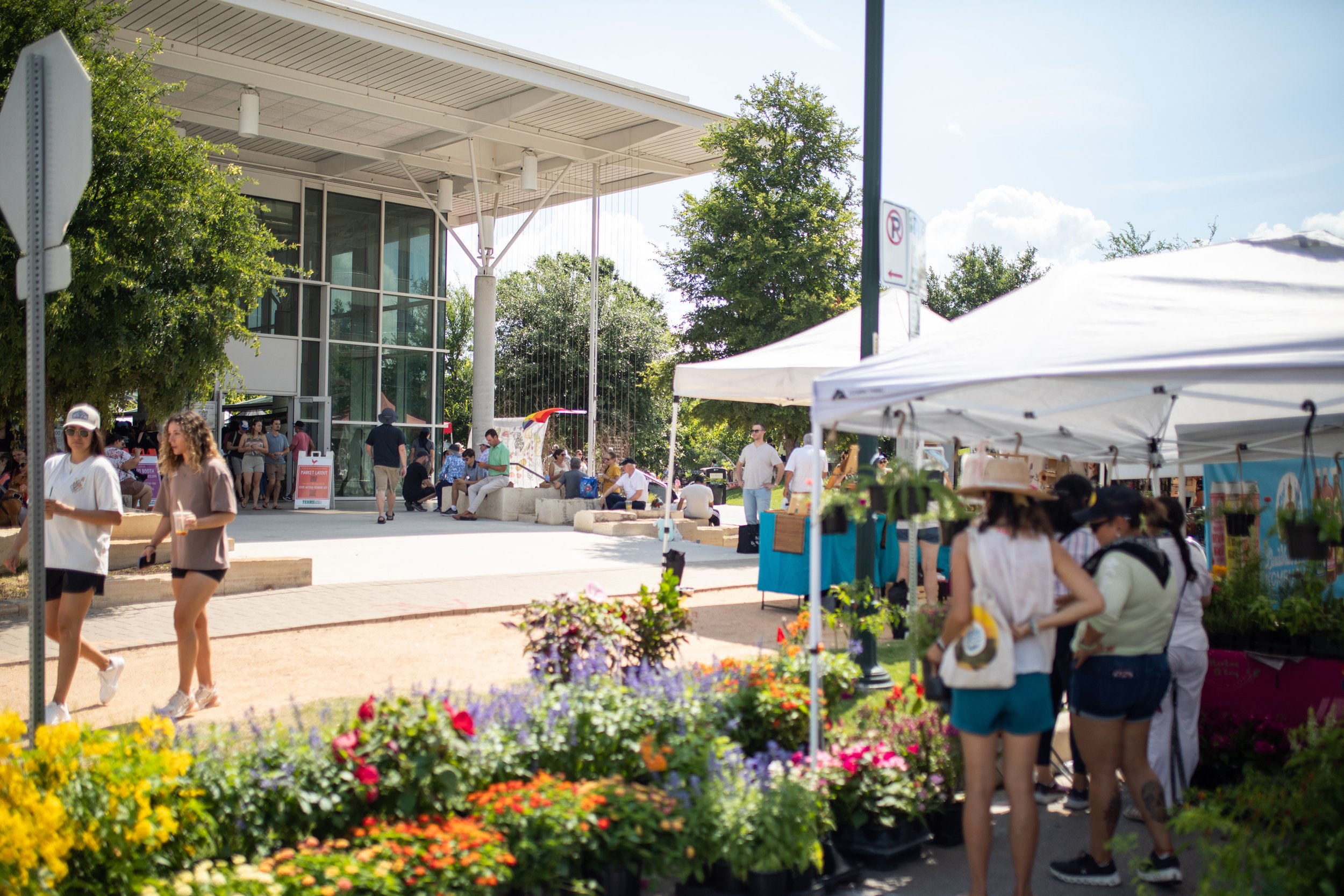 Outdoor market scene with people browsing stalls under white tents, colorful flowers in the foreground, and a modern glass building in the background.