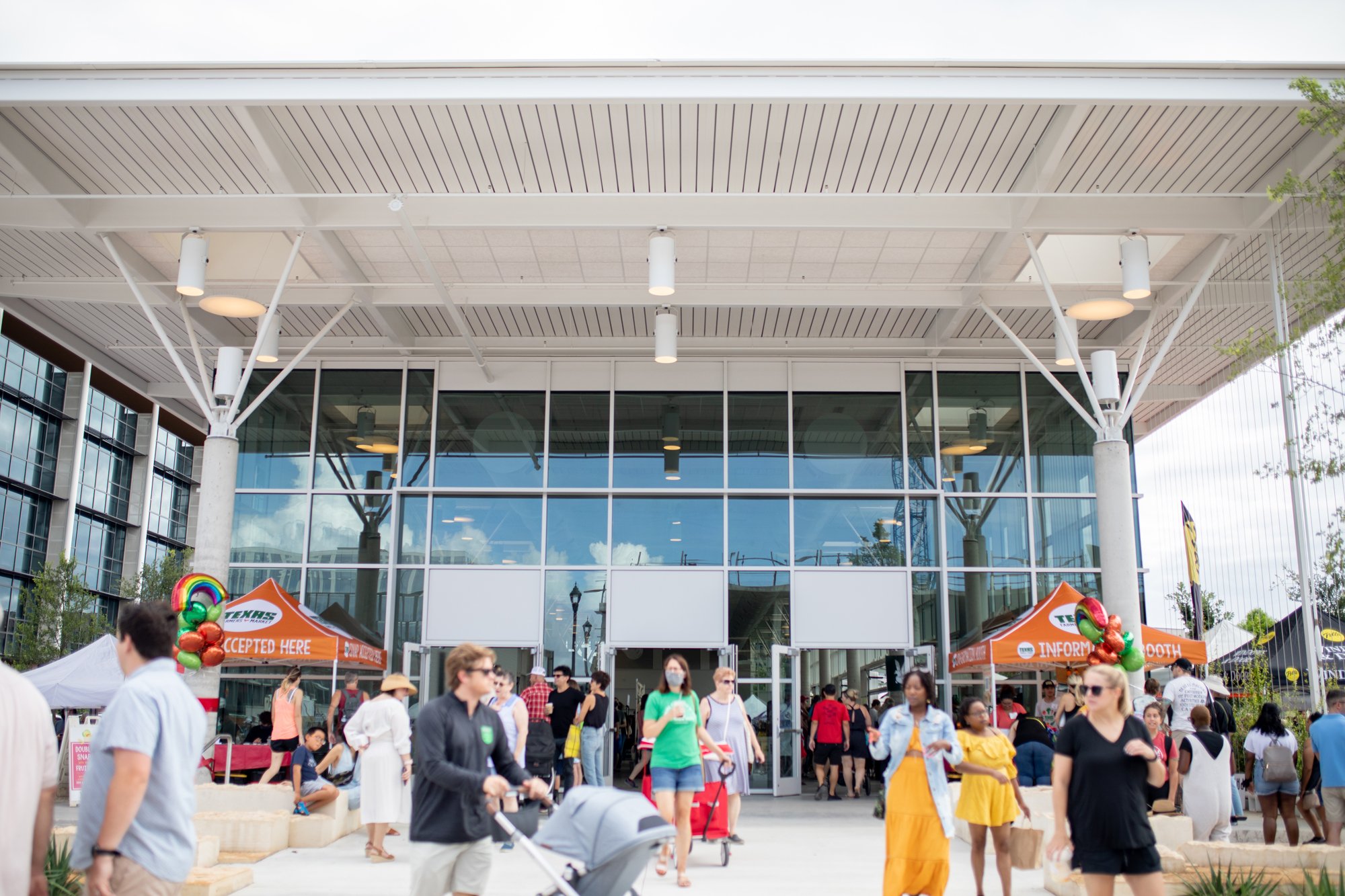 Crowd outside a modern building with glass windows, under a canopy, at a market event. People are walking with strollers and chatting. Vendor tents are visible, including an 'Information Booth' and a tent with a 'Cards Accepted Here' sign. Colorful b
