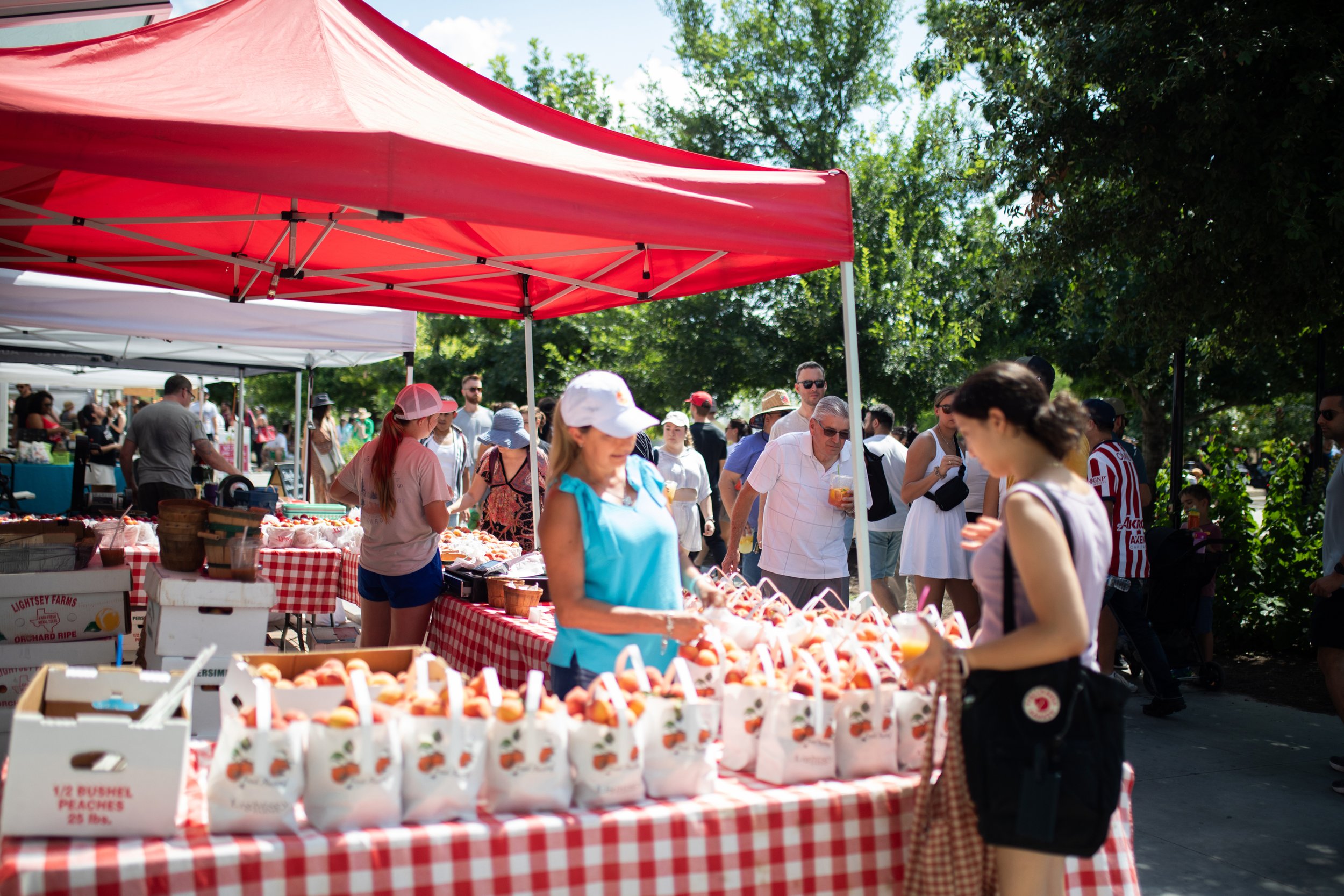 People shopping at a farmers market under red tents, with tables of fresh produce and a crowd in the background.