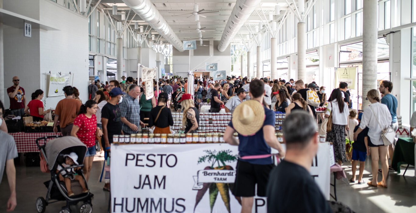 Indoor farmers market with people shopping, stalls selling jars of pesto, jam, and hummus, crowd of attendees, stroller with child, and visible signage.