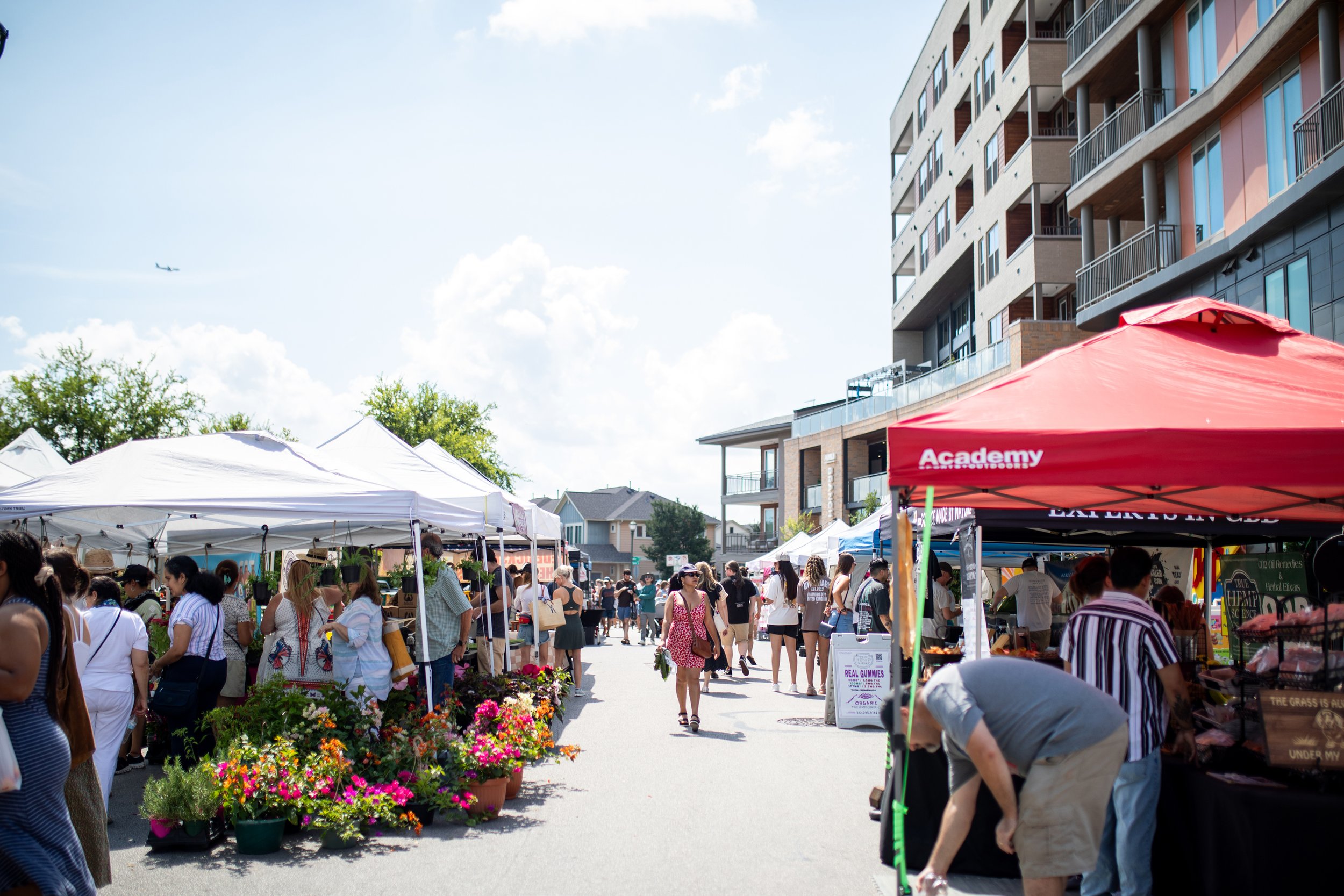 Outdoor farmers market with people shopping under vendor tents, colorful flowers on display, and apartment buildings in the background.