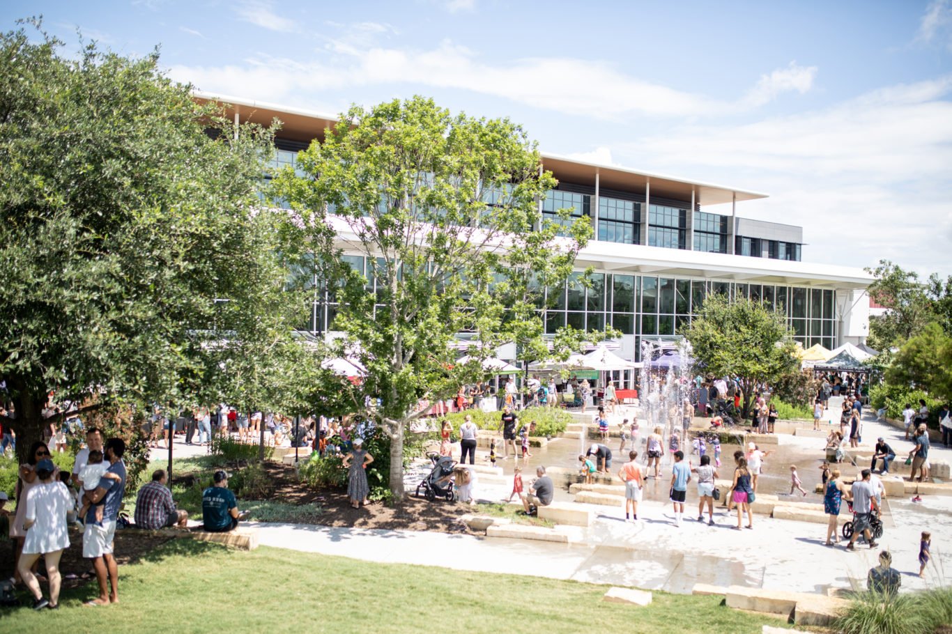 Outdoor event with people gathered around a water fountain, trees, and a modern building in the background on a sunny day.