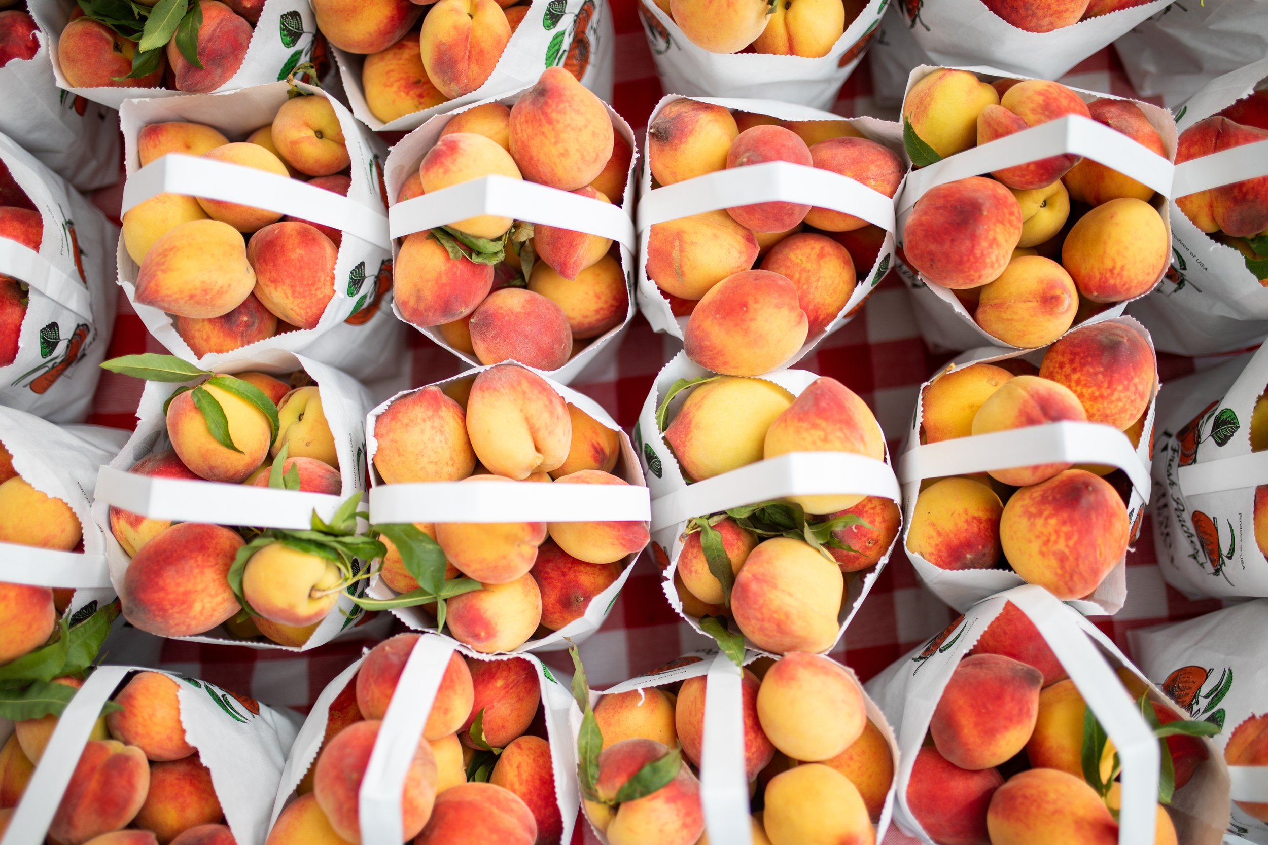 Paper baskets filled with fresh peaches on a checkered tablecloth.