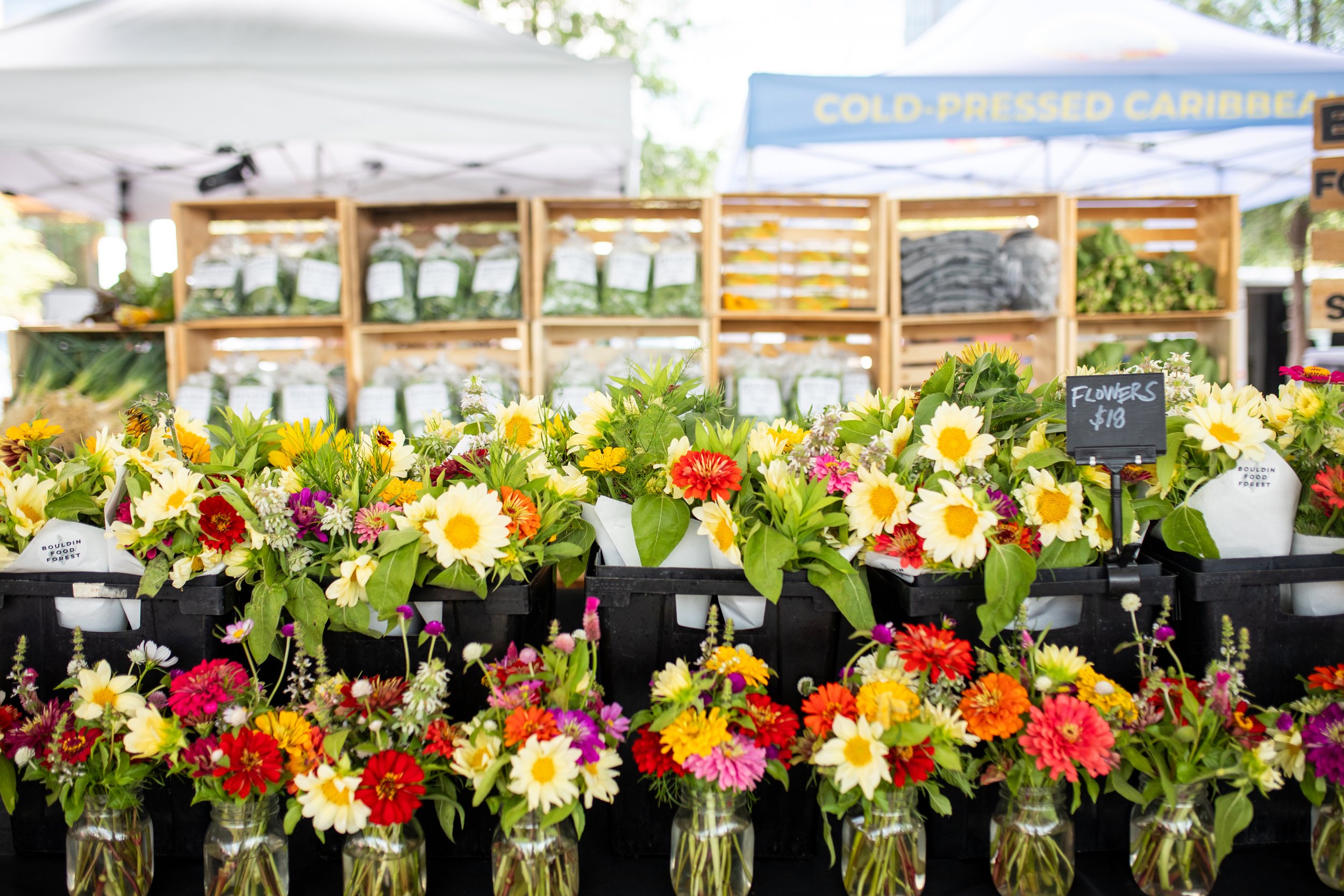 Colorful flower bouquets displayed at a farmers market with jars of mixed blooms in the foreground and wooden shelves with packaged greens in the background.