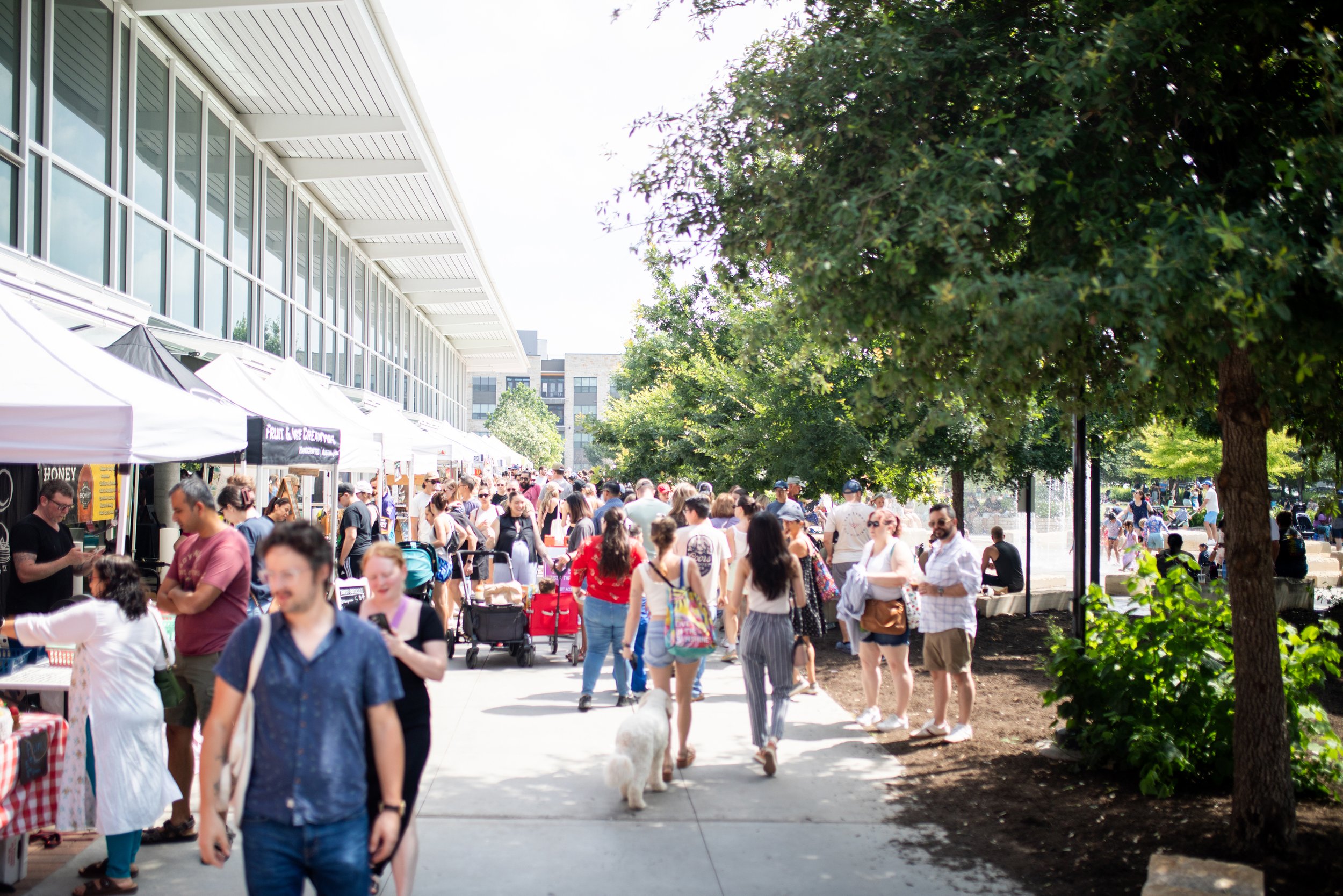 Crowded outdoor market with people walking past vendor stalls and trees nearby.
