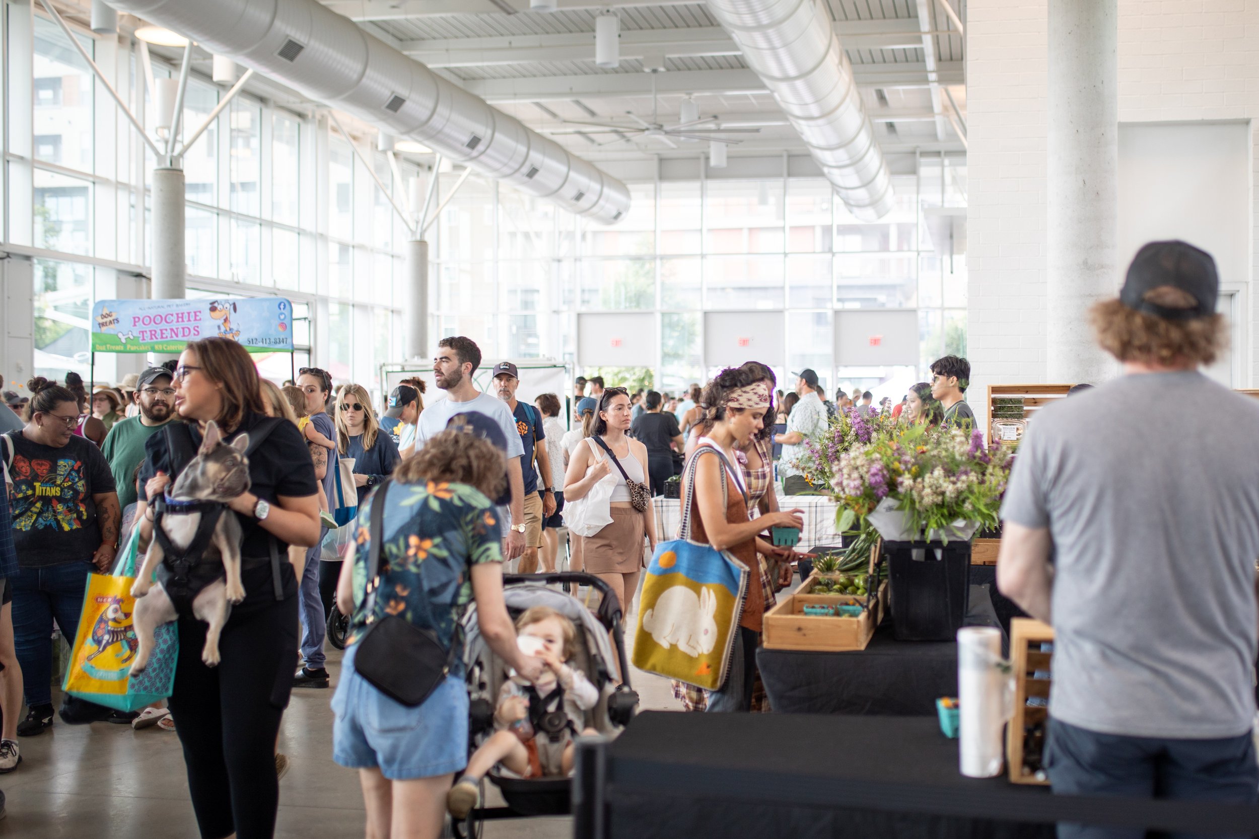 Indoor market with people shopping, holding tote bags, pushing a stroller, and interacting with various stalls. A woman carries a dog in a harness.