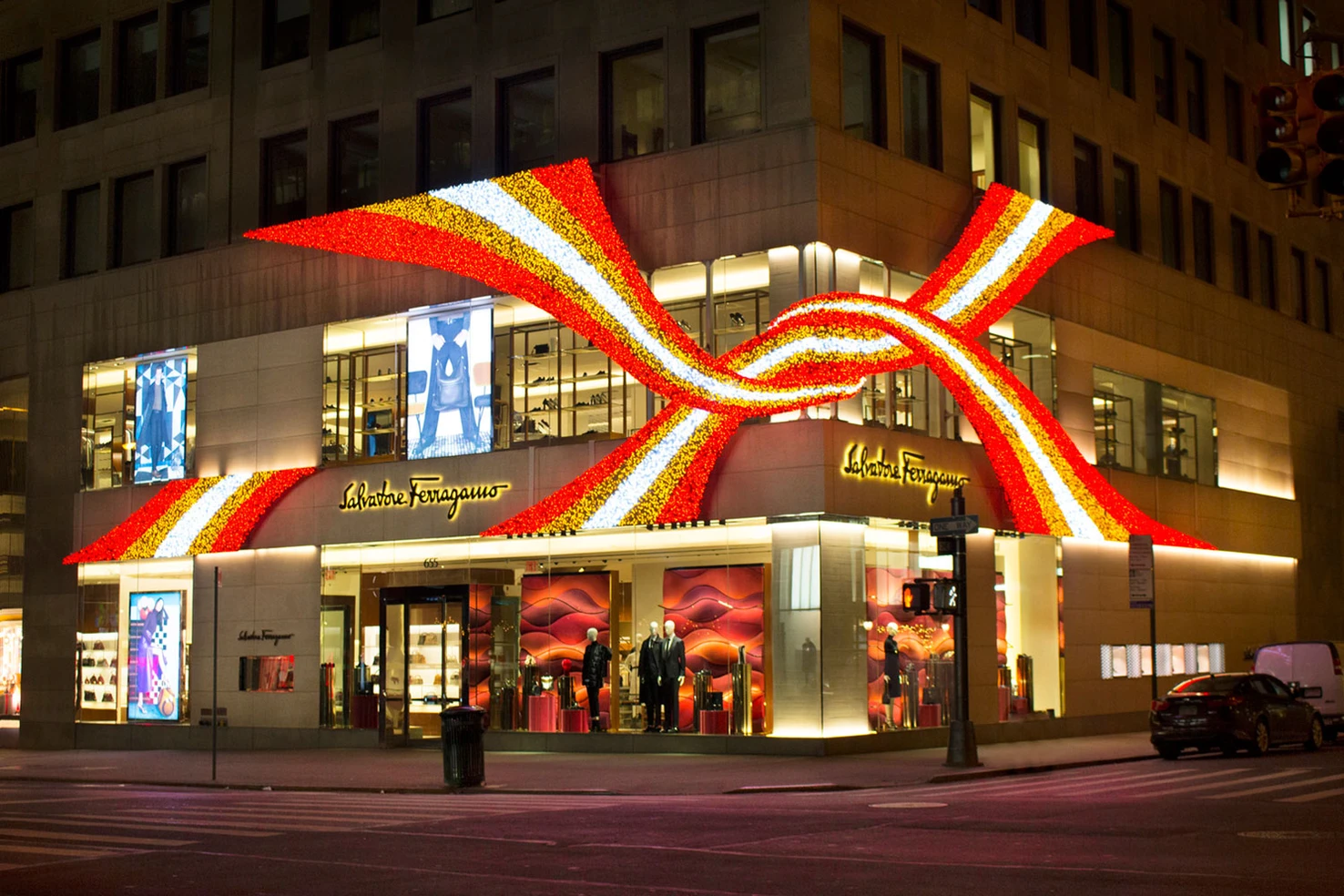 A Christmas or holiday display on a Saks Fifth Avenue store featuring a large, colorful bow made of red, yellow, and white lights. The store has mannequins inside and window displays with shoes and clothing.