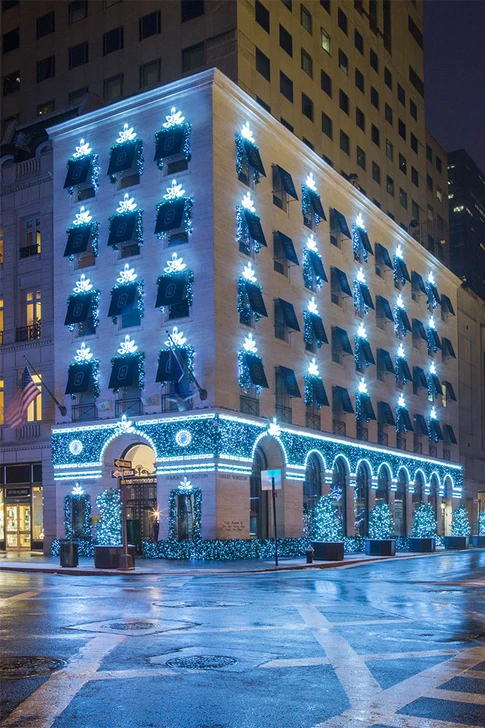 A multi-story building decorated with blue and white Christmas lights at night, featuring illuminated window displays and arched ground-floor windows.