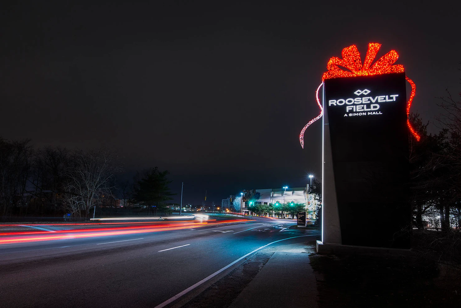 Nighttime view of Roosevelt Field shopping mall entrance with illuminated sign and holiday decorations, with light trails from passing cars on the road.