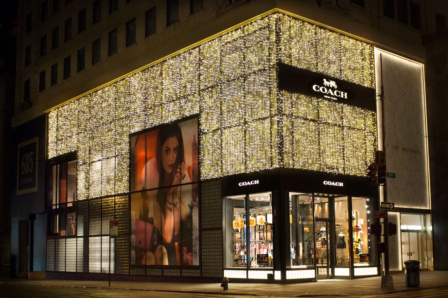 A nighttime view of a Coach store decorated with gold string lights covering the upper facade. The store has large glass windows displaying products, with a prominent advertisement featuring a woman holding a handbag. The Coach logo is visible above the entrance, and the building is located on a city street with a traffic light and street sign in the foreground.