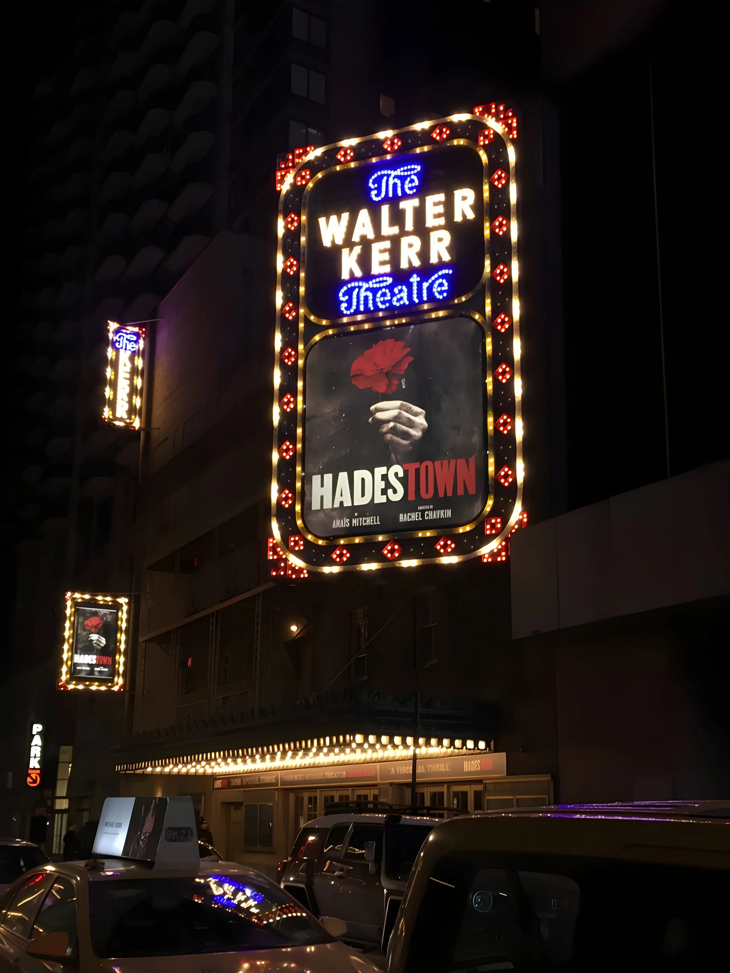 Nighttime view of the Walter Kerr Theatre marquee with lights, advertising the play 'Hades Town' with a poster showing a hand holding a red flower.