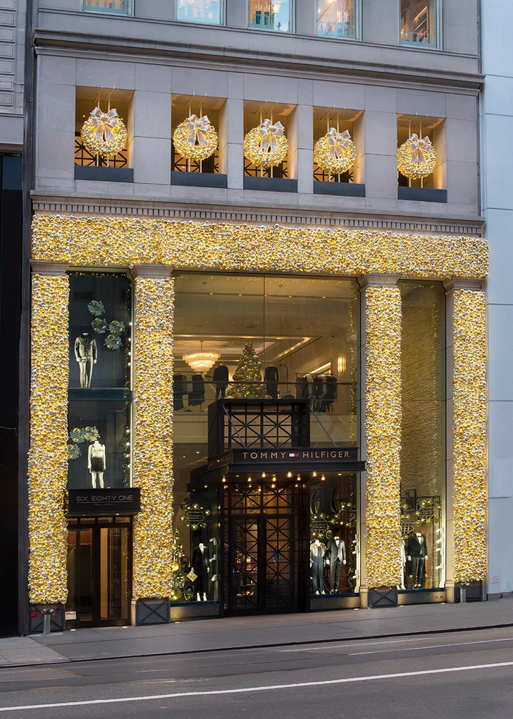 Storefront decorated with Christmas wreaths and floral arrangements, featuring mannequins dressed in formal clothing, and illuminated holiday decorations, including wreaths with bows and lights.