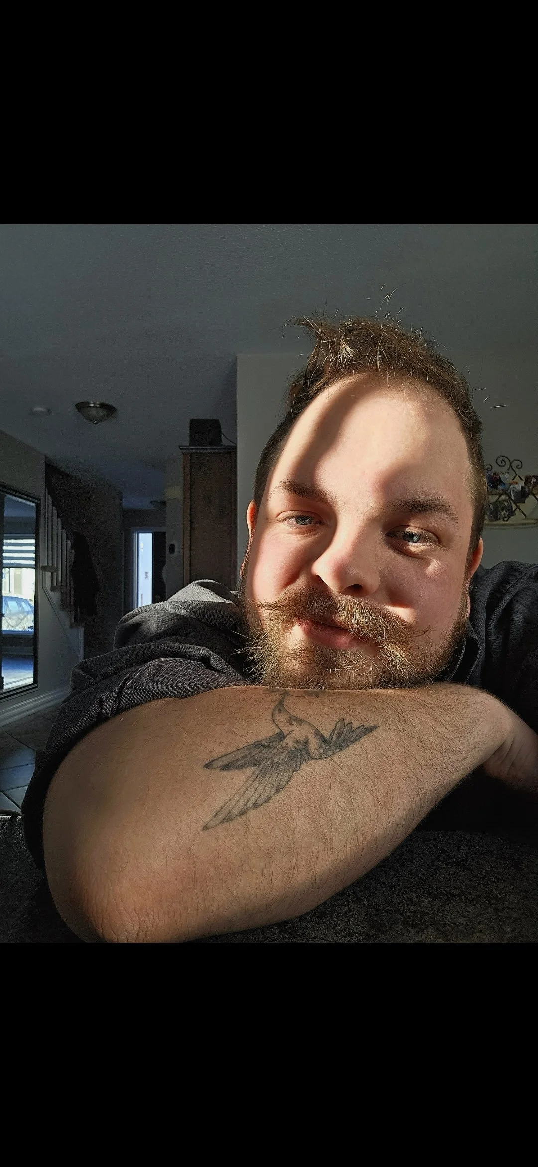 A man with a beard and mustache resting his head on his arm, which has a bird tattoo, in a room with natural sunlight.
