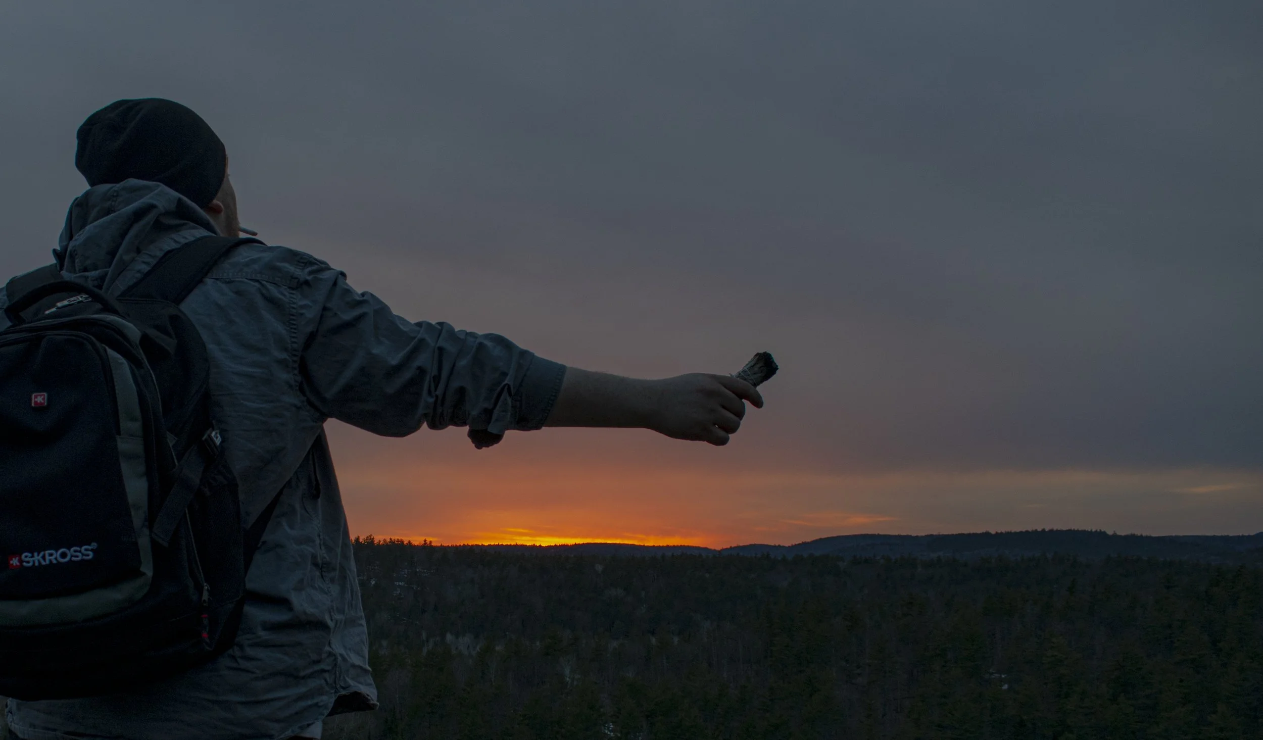 Person wearing a backpack and hoodie reaching out with a small sage stick during sunset over a forested landscape.
