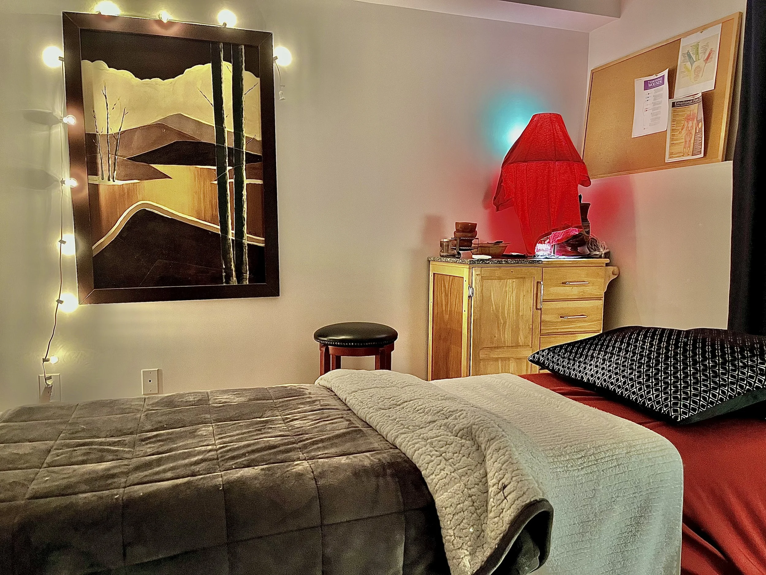 A bedroom with a bed covered in a brown quilt and a cream blanket, a black and white patterned pillow, a wooden dresser topped with a red lampshade, a stool, a framed landscape painting, and a bulletin board on the wall.