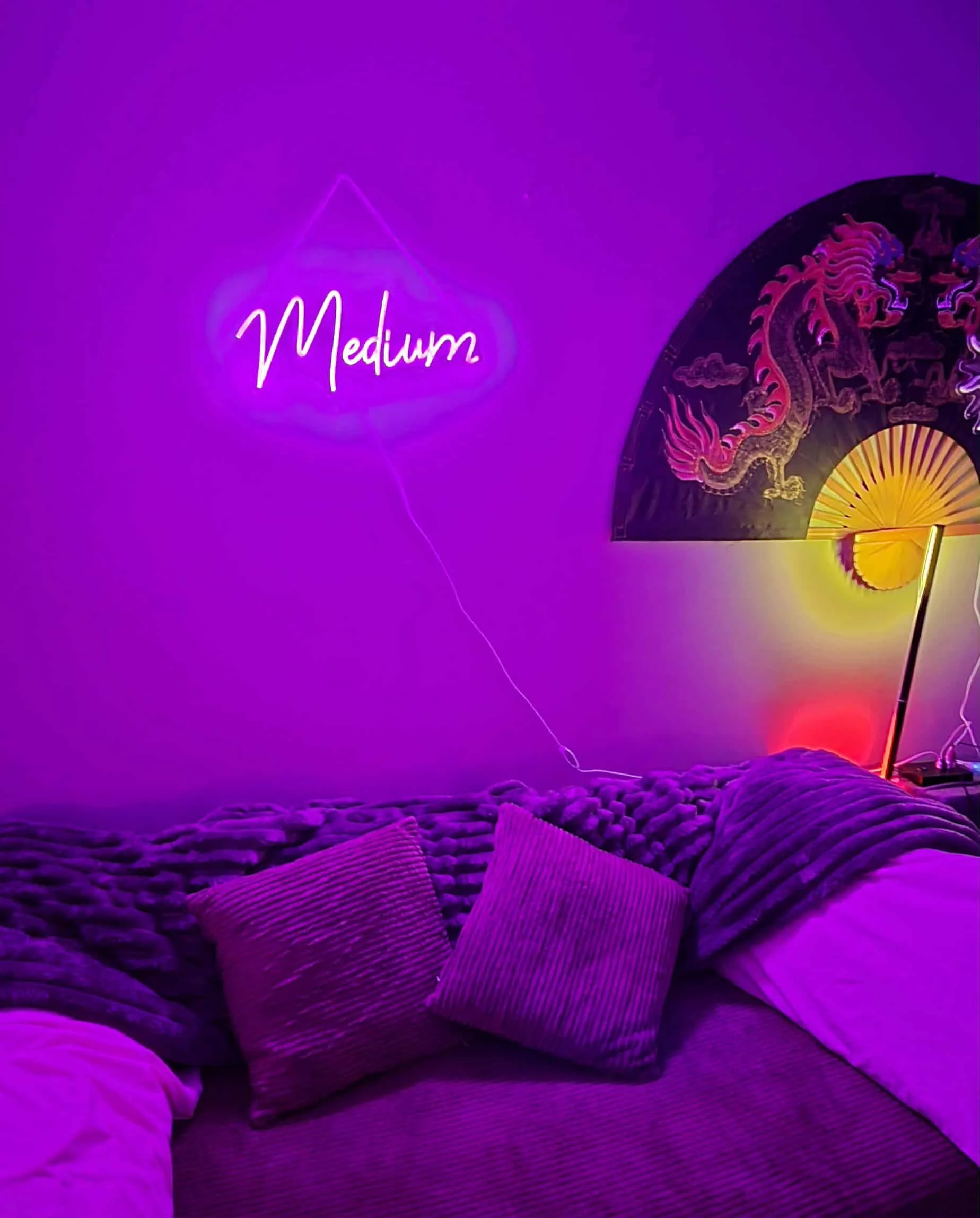 A room with purple ambient lighting, a neon sign that says 'Medium', a decorative folding fan with a dragon design, and purple pillows on a textured couch.