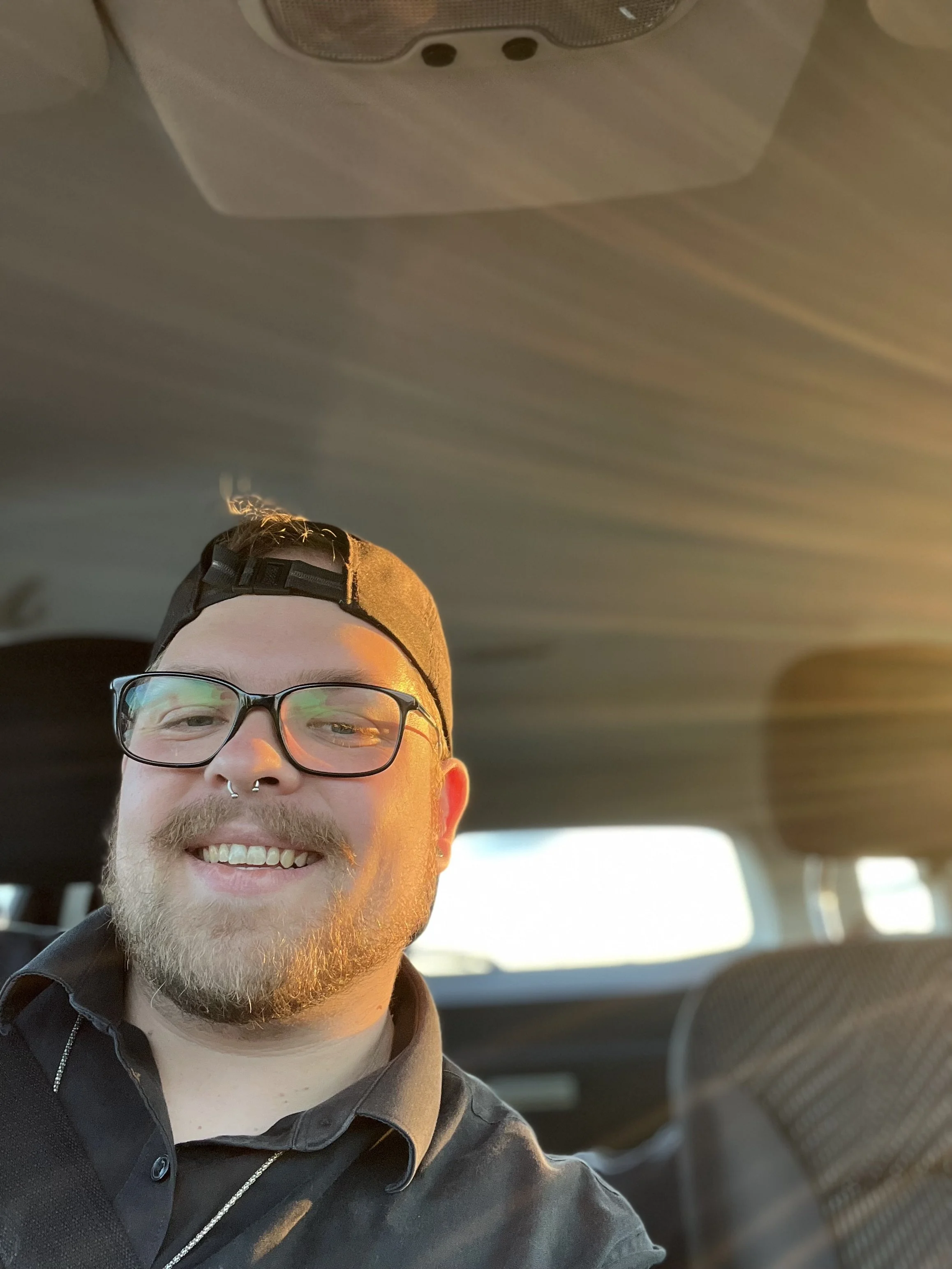 A young man with glasses, a septum piercing, and a beard is smiling in the backseat of a car. The photo is taken during sunset or sunrise, casting a warm glow on his face.