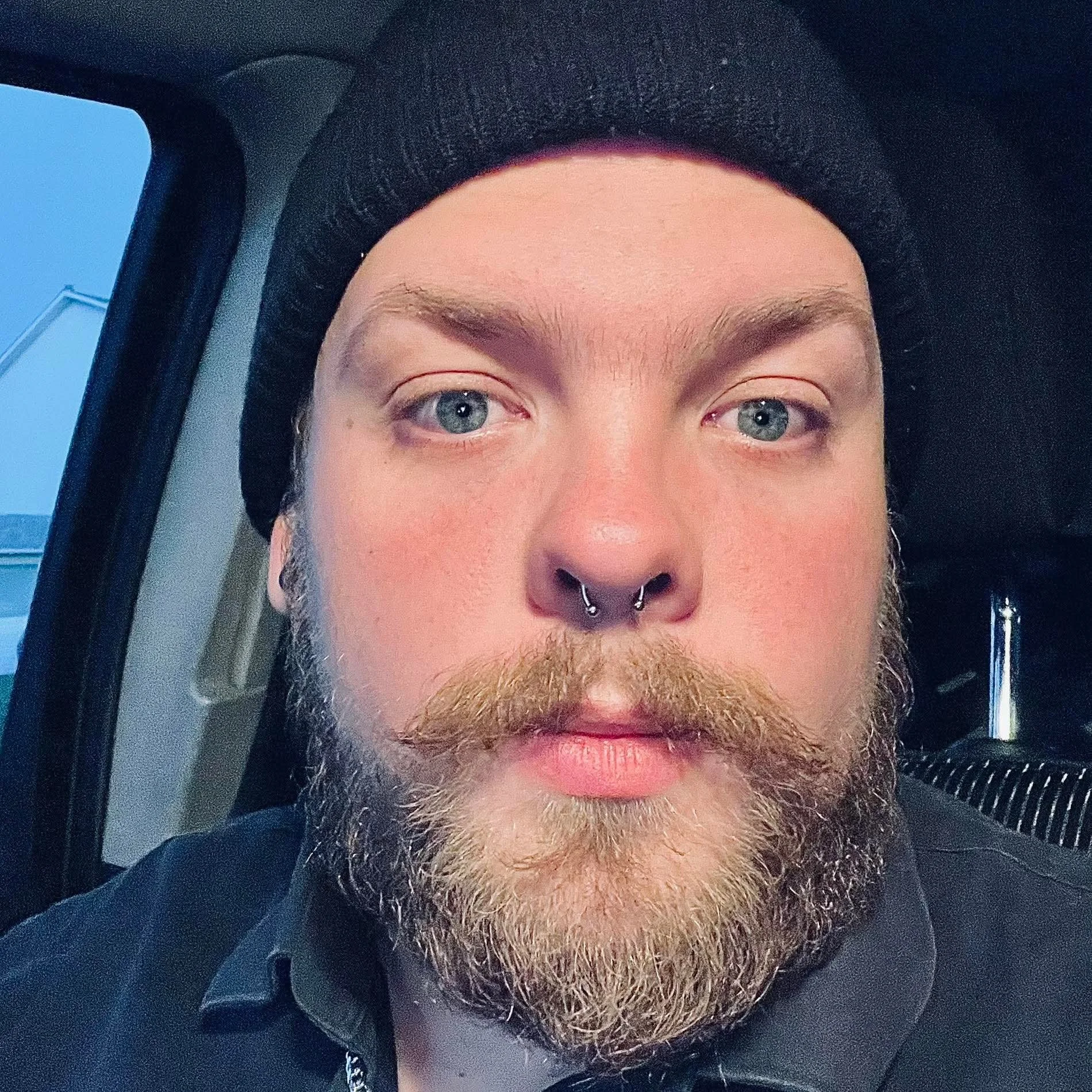 Close-up selfie of a young man with blue eyes, a beard, a septum piercing, wearing a black beanie, and a black shirt. He is inside a vehicle, with a window and part of the interior visible.