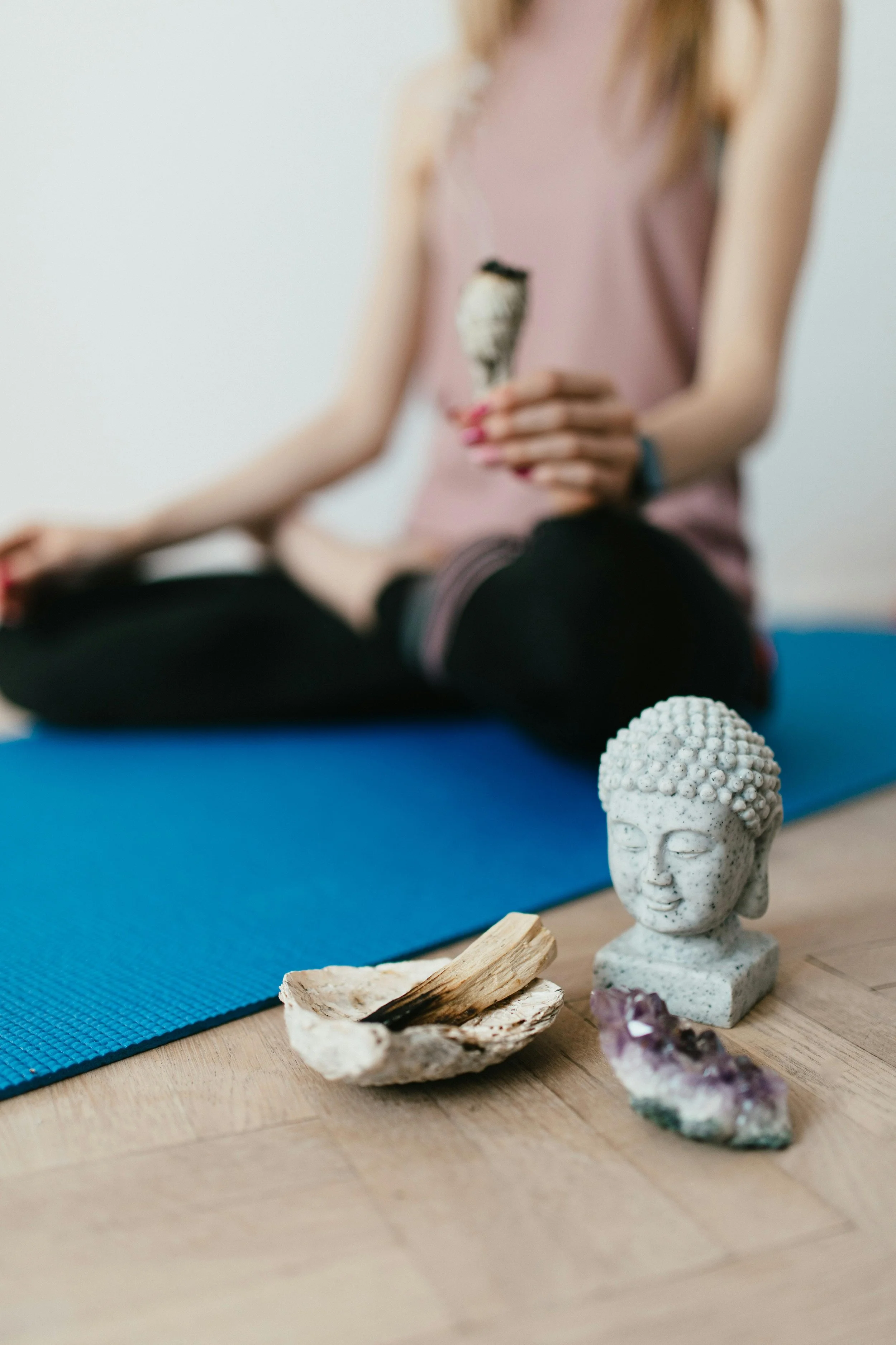 Close-up of a yoga meditation space with a Buddha head sculpture, a small amethyst crystal, and a bowl with wood pieces, with a woman sitting cross-legged on a mat in the background.
