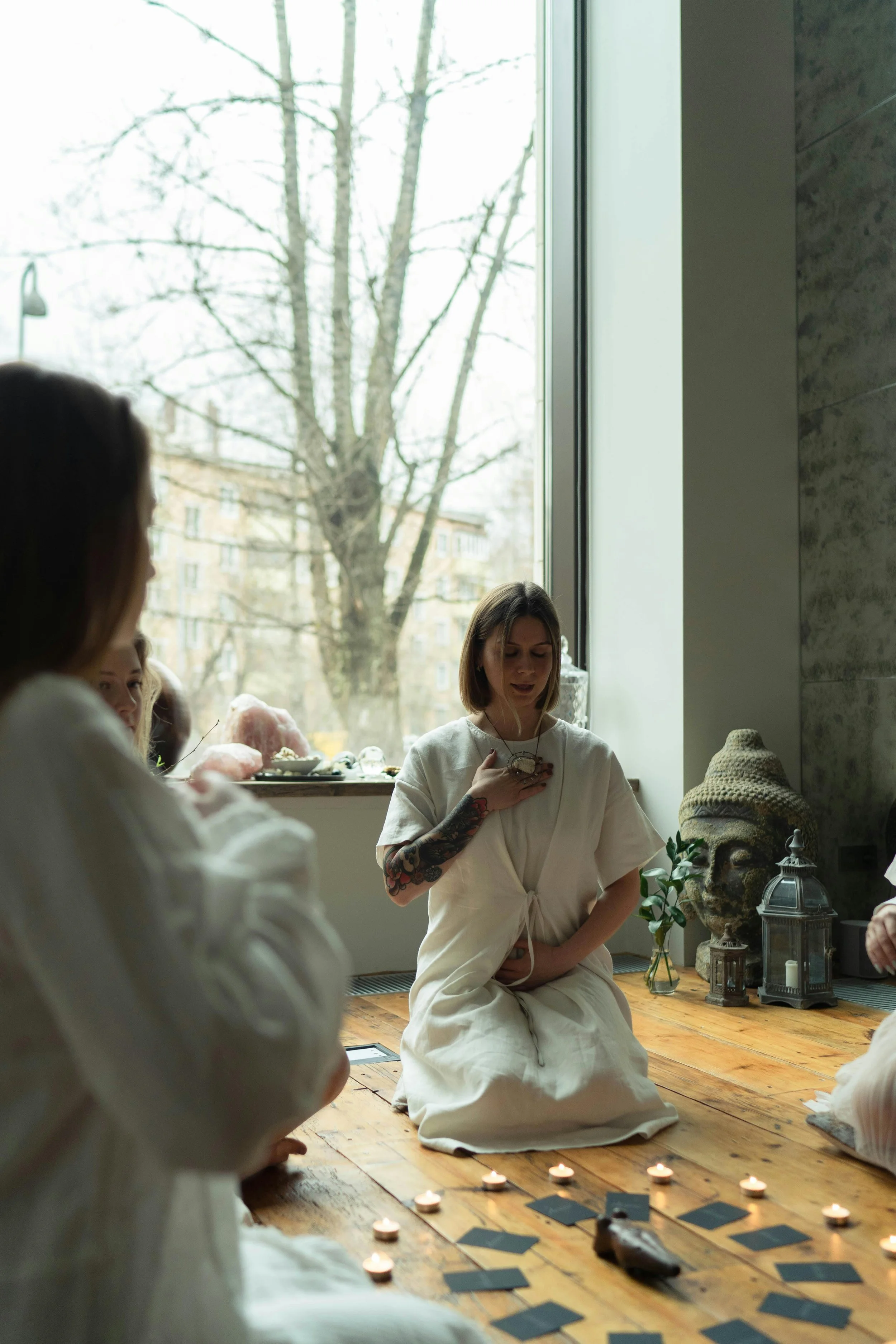 A group of people participating in a spiritual or meditation session in a room with large windows showing trees outside. One woman is meditating with her eyes closed, hands on her chest, and other participants are sitting around her.