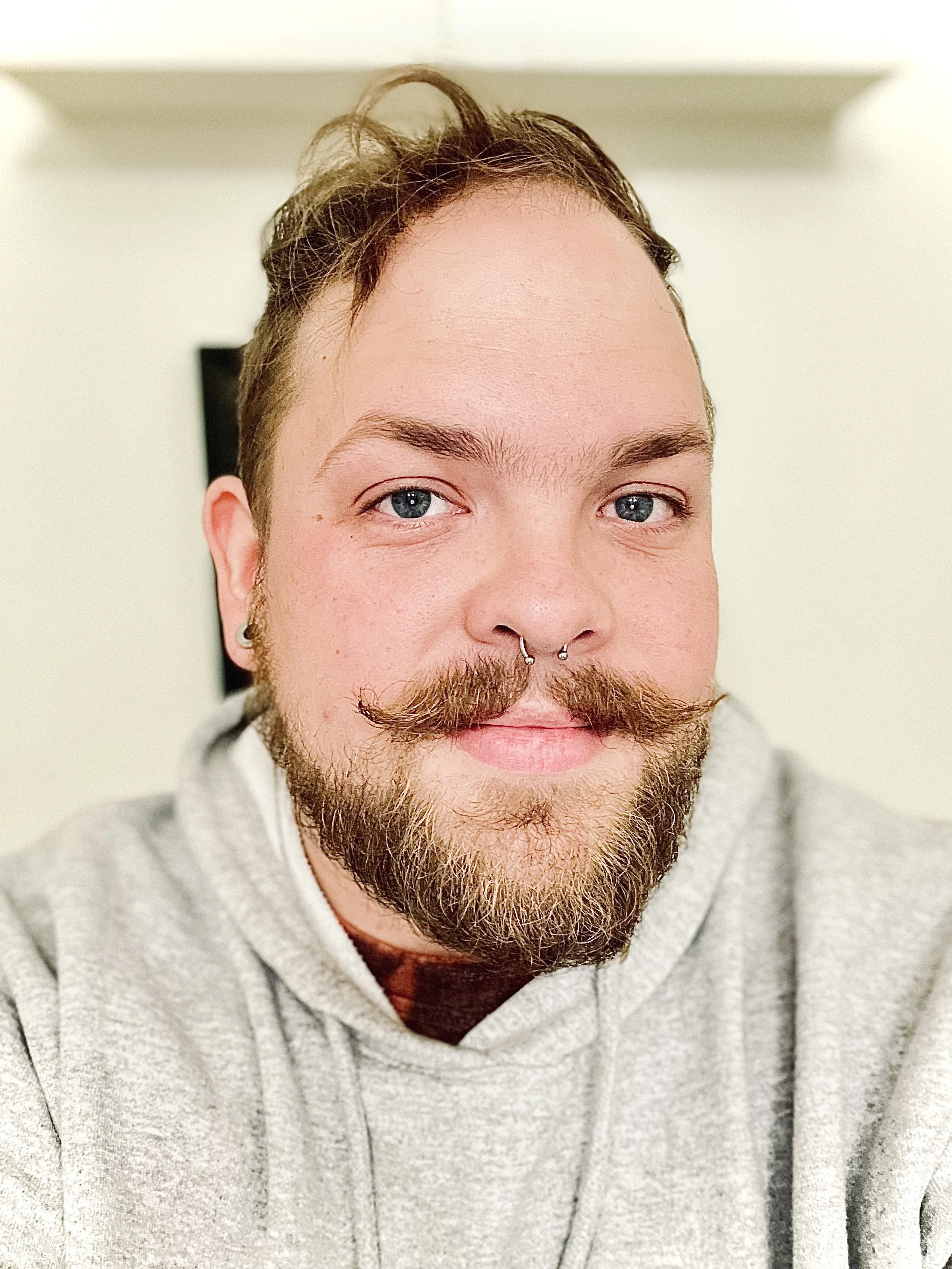 Close-up photo of a young man with blue eyes, a beard, mustache, and a septum piercing, wearing a gray hoodie and smiling slightly.