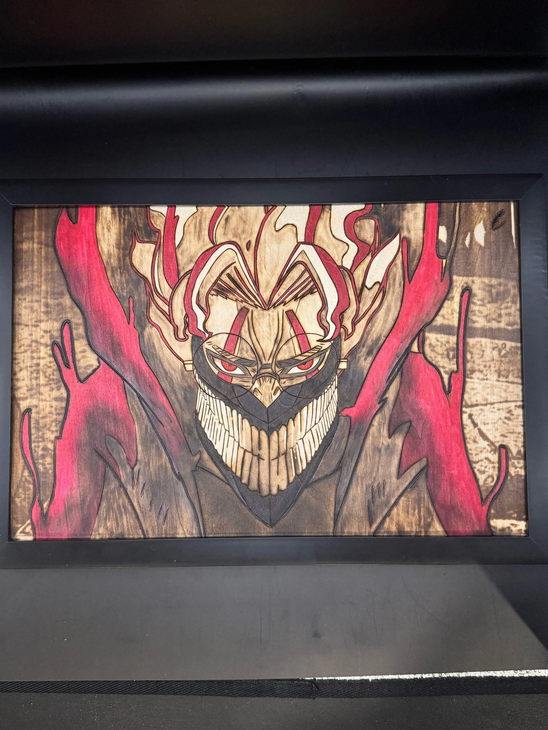 A framed artwork of a menacing character with sharp teeth and glowing red eyes, surrounded by red and black flames, on a wooden background.