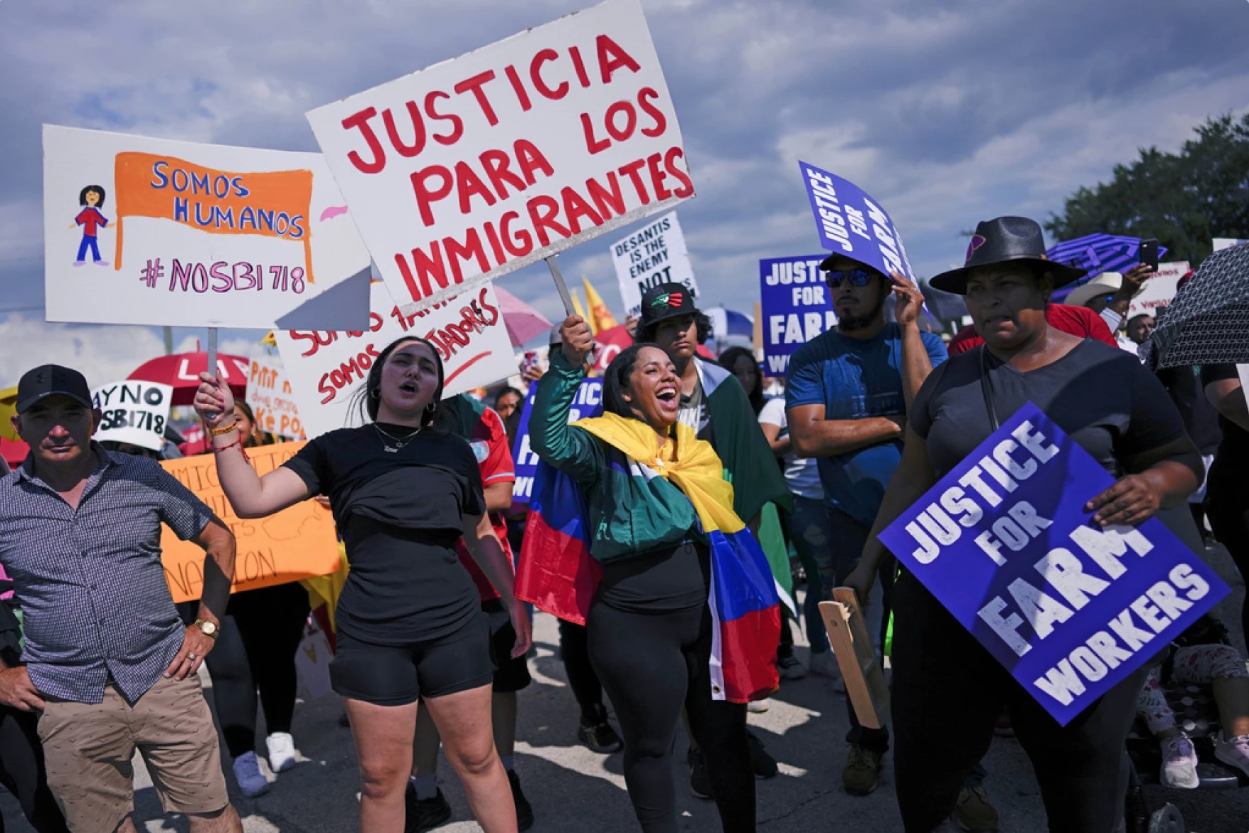A crowd of people protesting for immigrant and worker justice, holding signs with messages like 'Justice for Immigrants' and 'Justice for Farm Workers', in an outdoor setting with a blue sky and clouds.