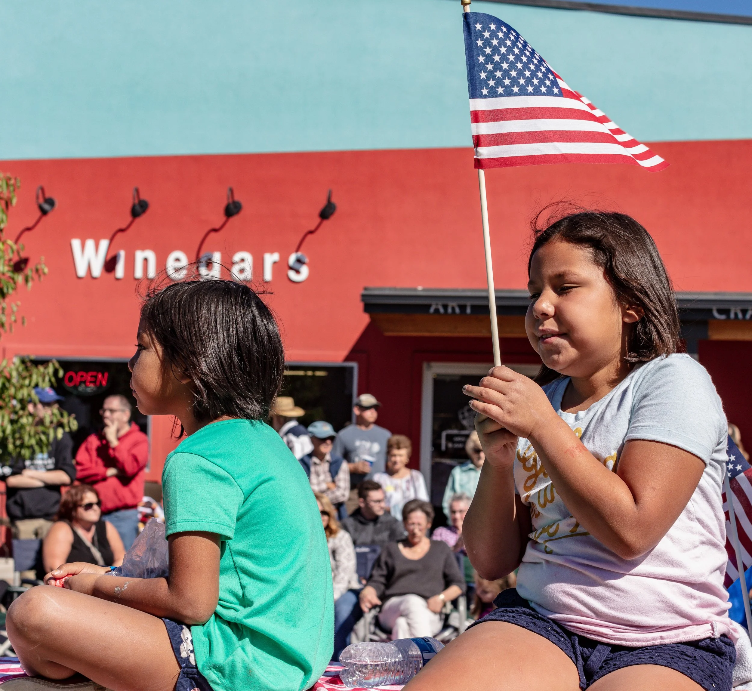 Two young girls sitting on the ground during a parade, one holding an American flag, with a crowd of people watching in front of a red building with the sign 'Winedars.'