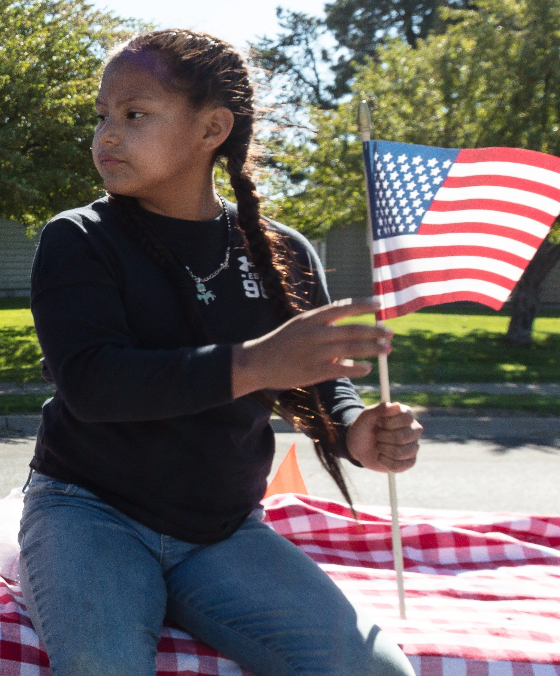 A young girl sitting on a red and white checkered blanket, holding an American flag outdoors with trees and houses in the background.