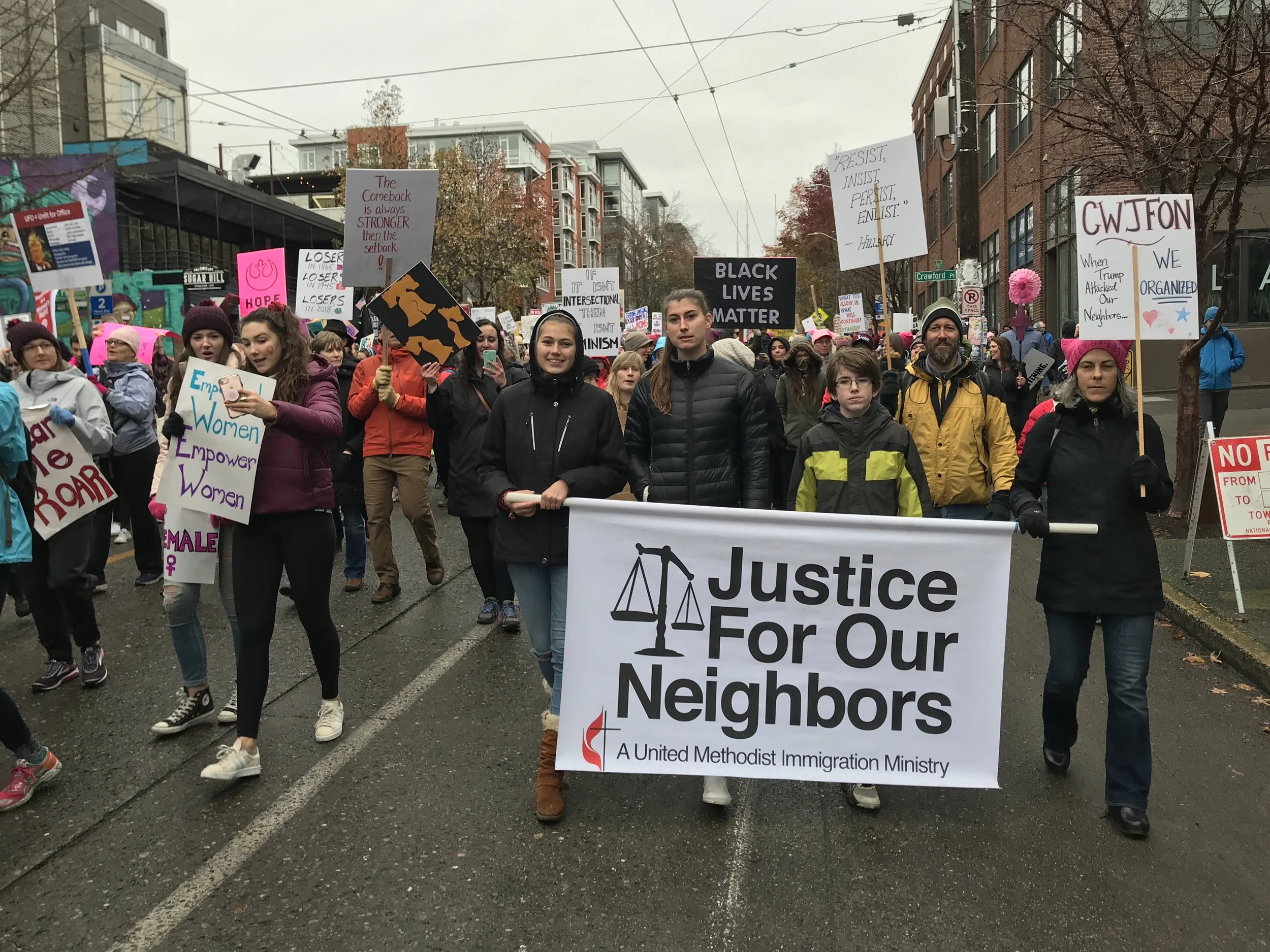 A diverse group of people participating in a protest or march on a city street, holding signs advocating for social justice, equality, and immigrant rights. The main sign reads 'Justice For Our Neighbors' and others include messages like 'Black Lives Matter' and 'Women Empower Women'.