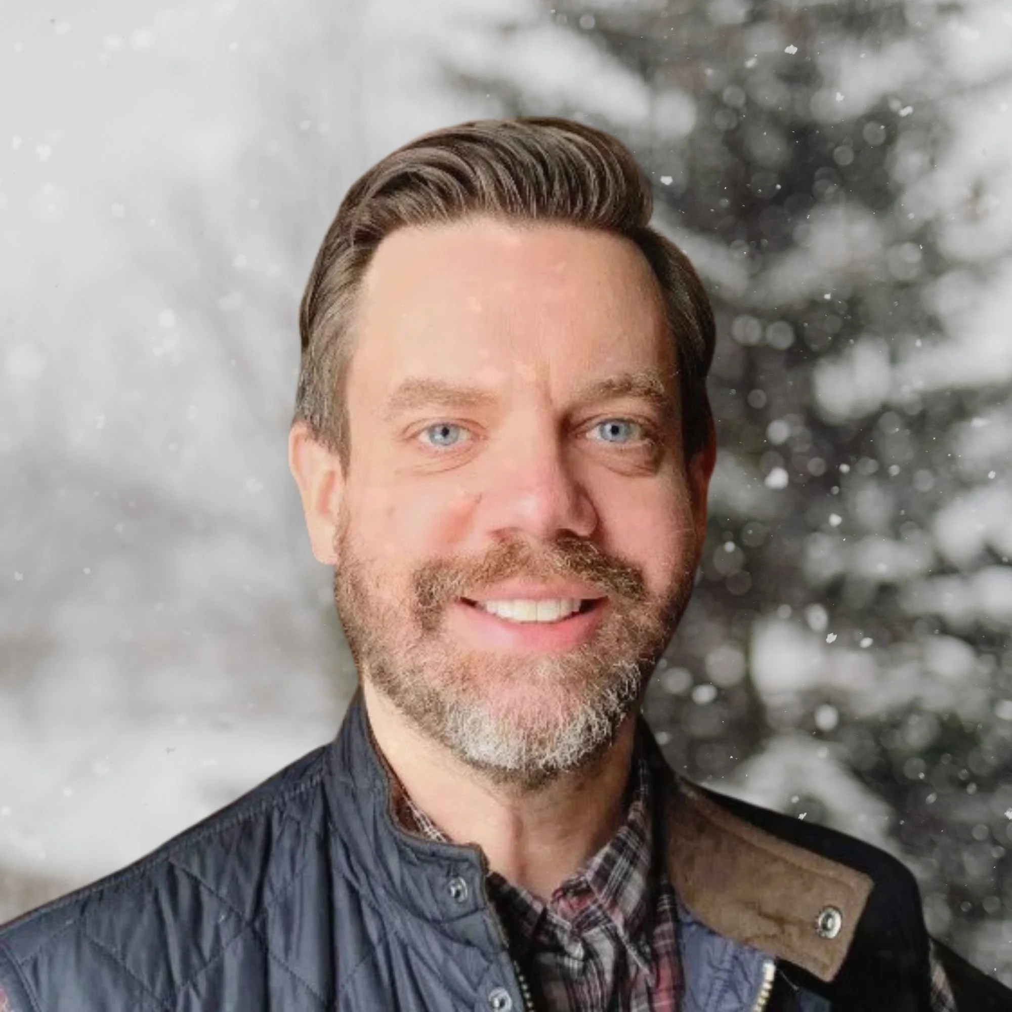 A smiling man with light brown hair, a beard, and blue eyes, dressed in a plaid shirt and a blue quilted jacket.