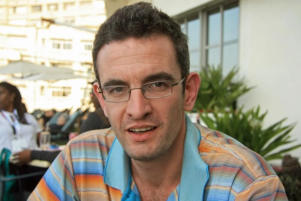A man with glasses and curly hair wearing a multicolored striped shirt, sitting outdoors at a restaurant table with other people and greenery in the background.