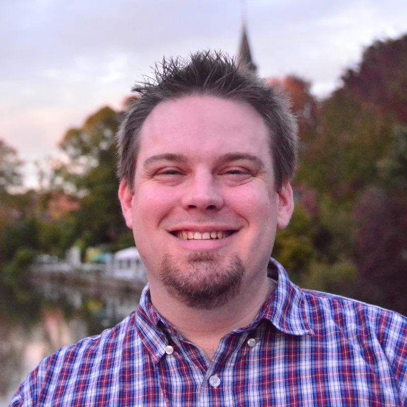 A smiling man with spiked hair and facial hair, wearing a plaid shirt, standing outdoors in front of a river with trees and a church steeple in the background during sunset.