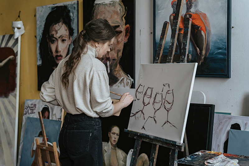 Artist working in a studio, developing an original painting surrounded by finished artworks and works in progress.