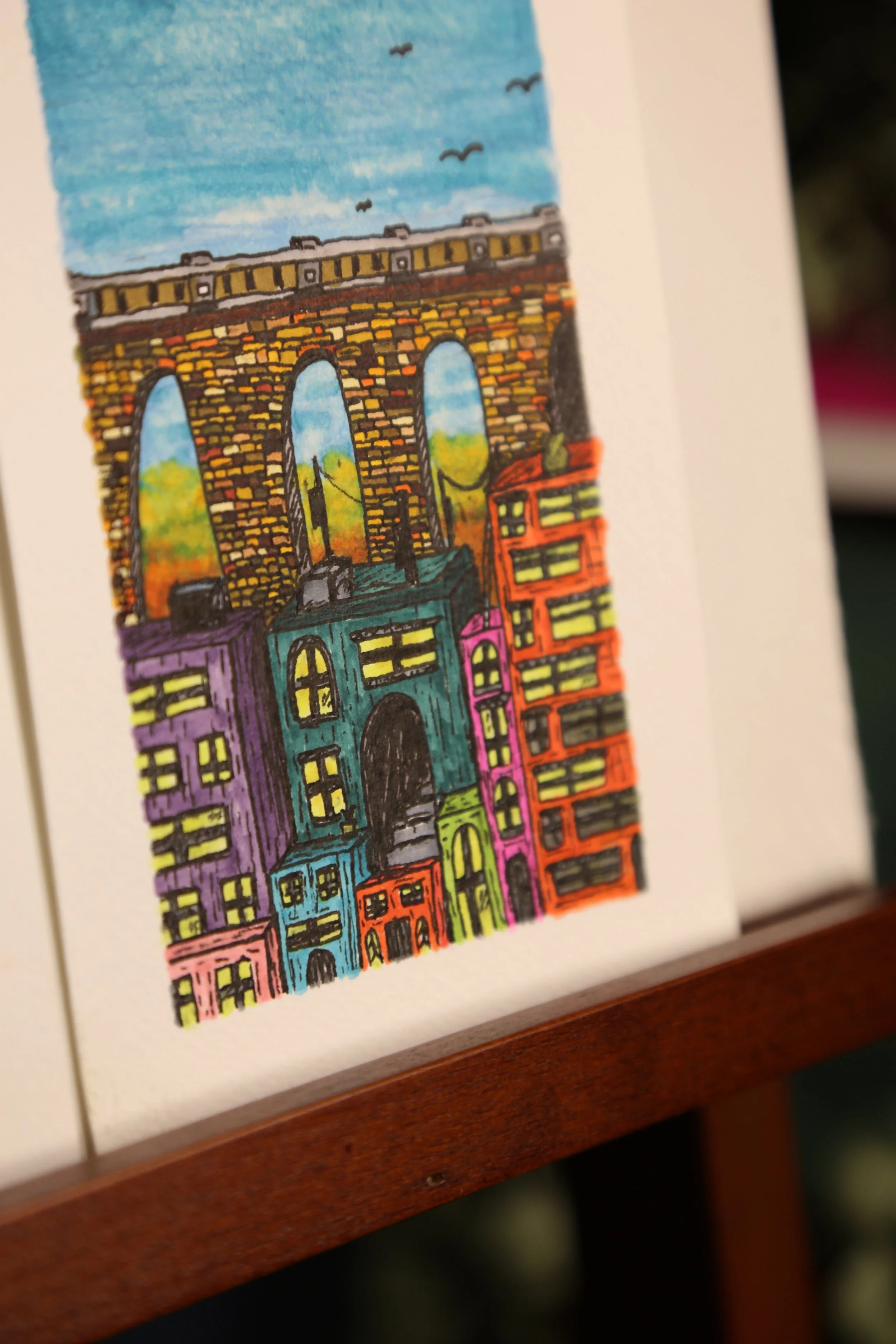 Close-up corner detail of Bridge Scene by Rix, showing colorful hand-drawn buildings beneath a stone arch bridge on watercolor paper.