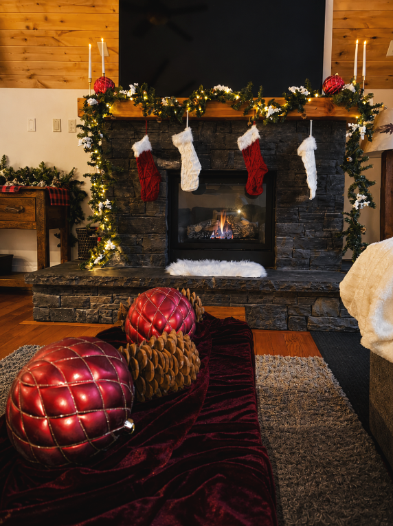 Living room with a stone fireplace decorated for Christmas, featuring four stockings hanging from the mantel, illuminated candles, greenery, and Christmas ornaments. There is a red velvet runner on the floor with large red and pinecone decorations, and a cozy scene with a lit fireplace.